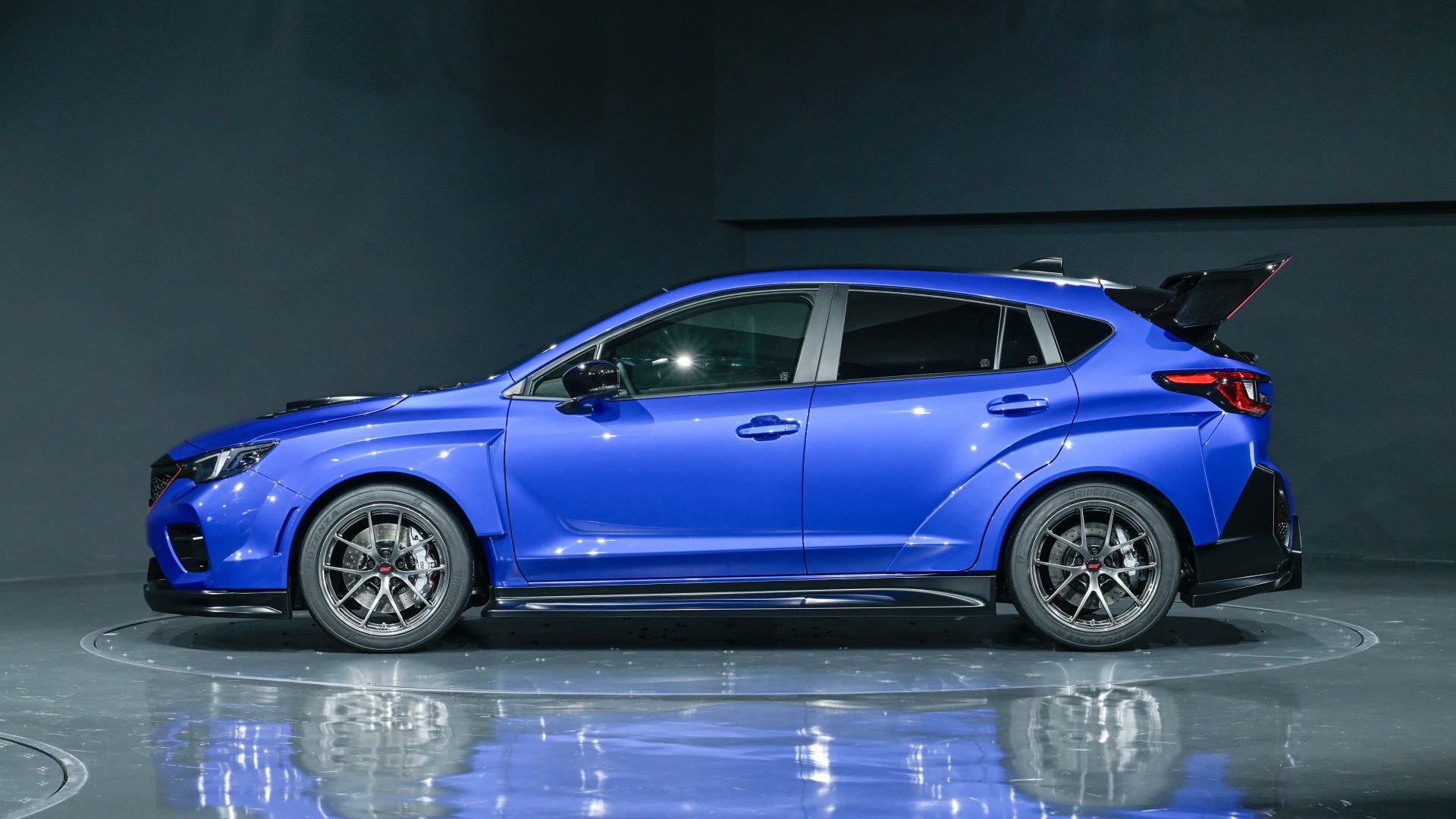 Blue sporty hatchback car with black alloy wheels and a large rear spoiler in a studio setting.
