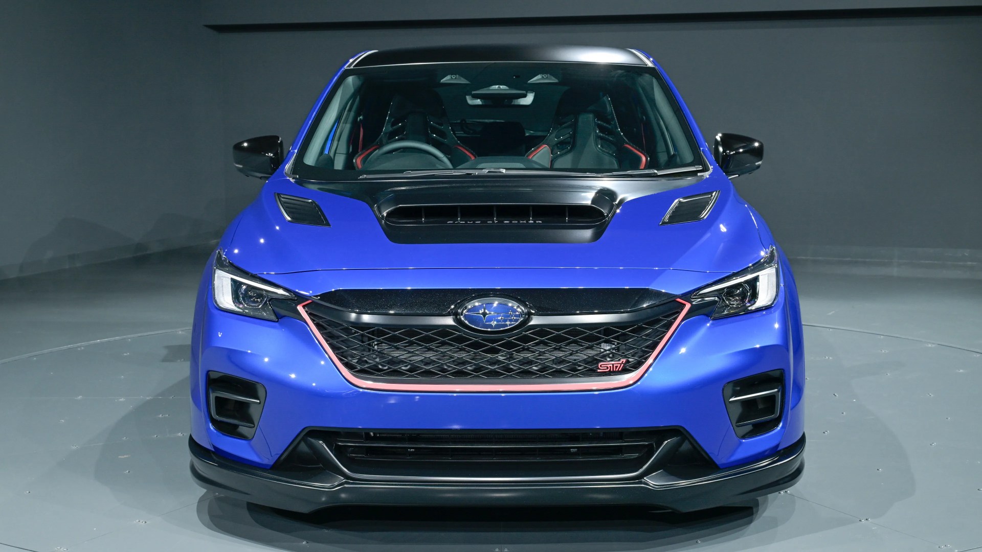 Blue Subaru STI car front view with black hood scoop and red trim on grille.