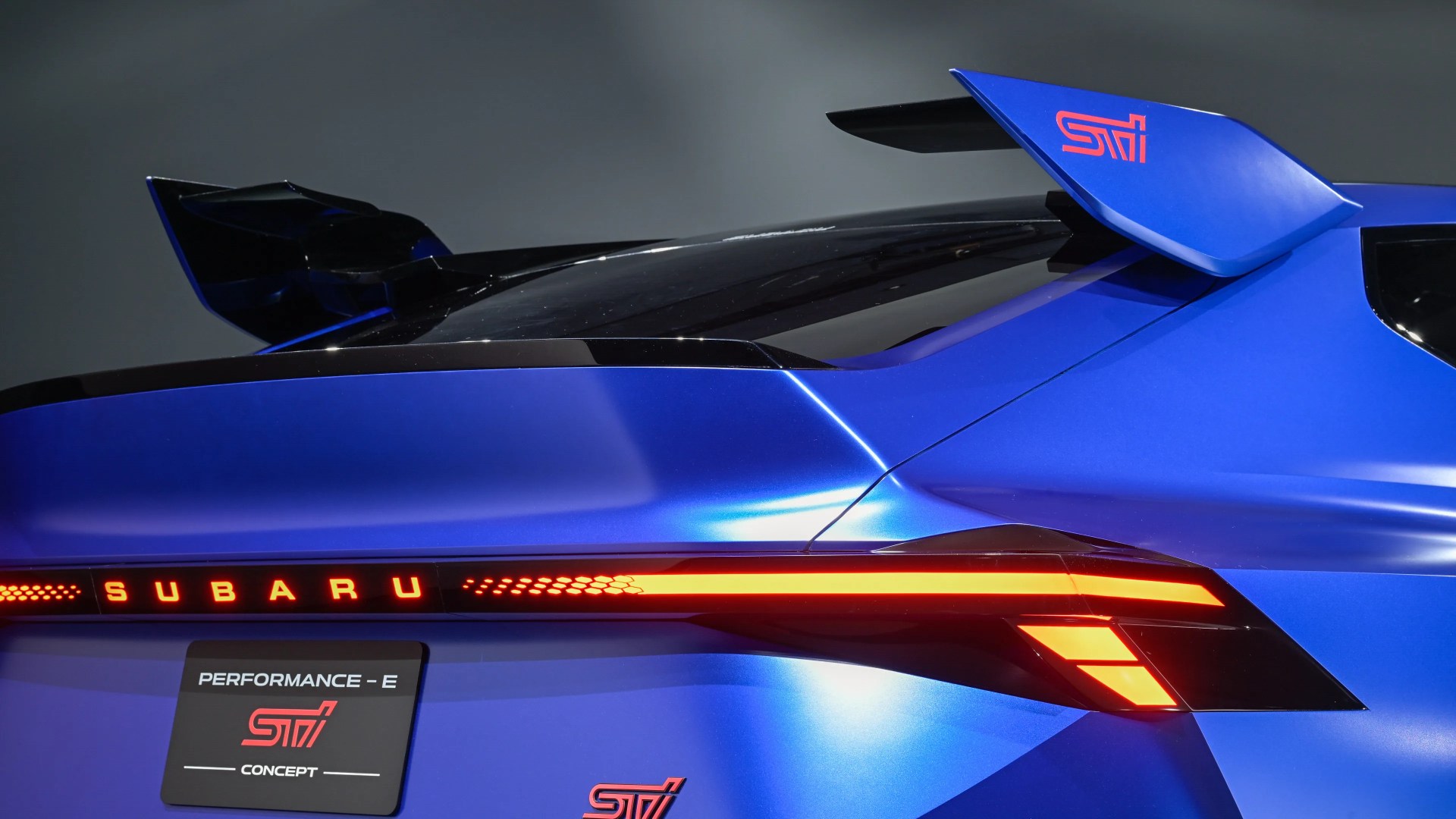Rear side of a blue Subaru Performance-E STI concept car with illuminated red taillights and a large rear spoiler.