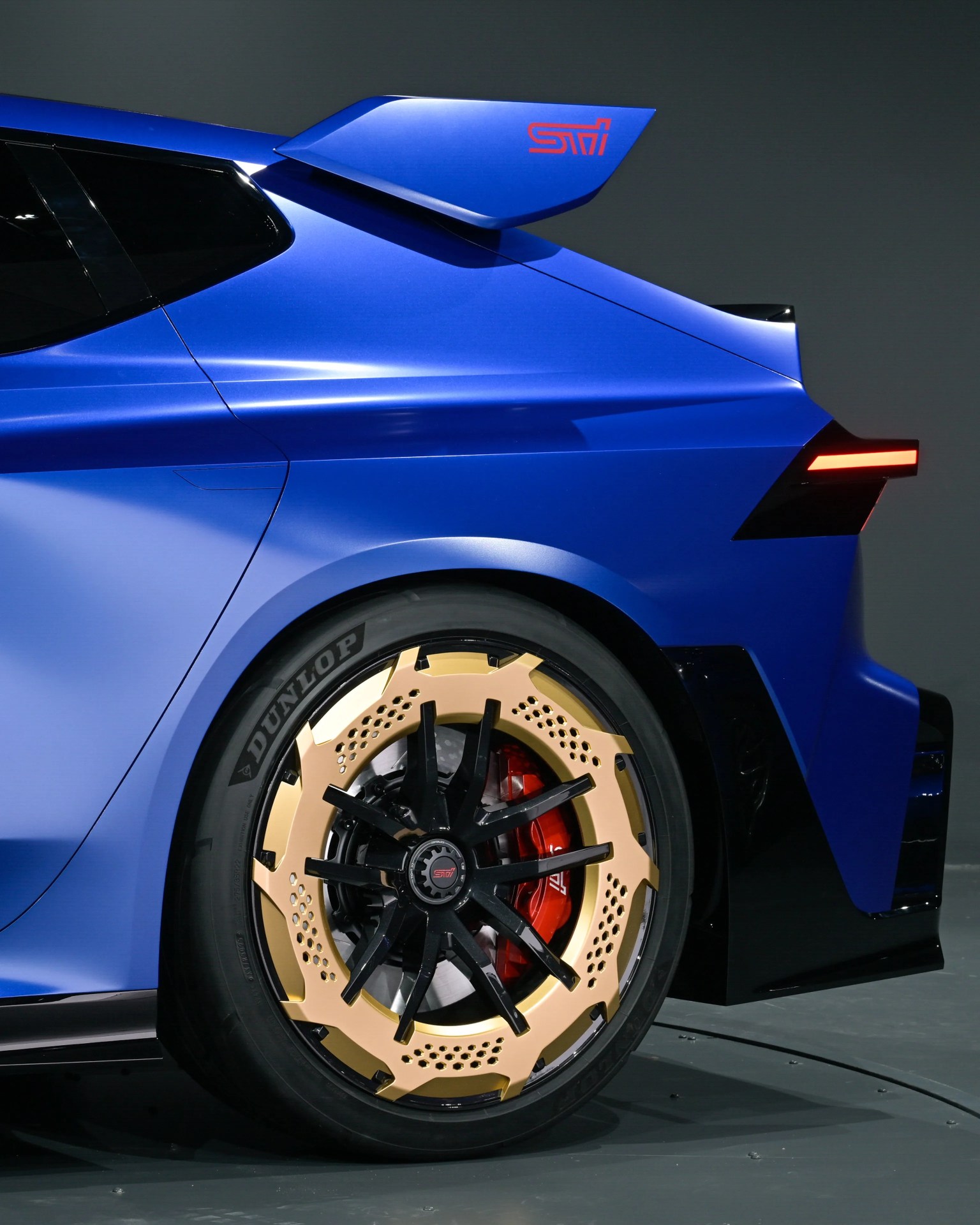 Rear side of a blue car with a large rear wing, black and gold multi-spoke wheel, and red brake caliper.