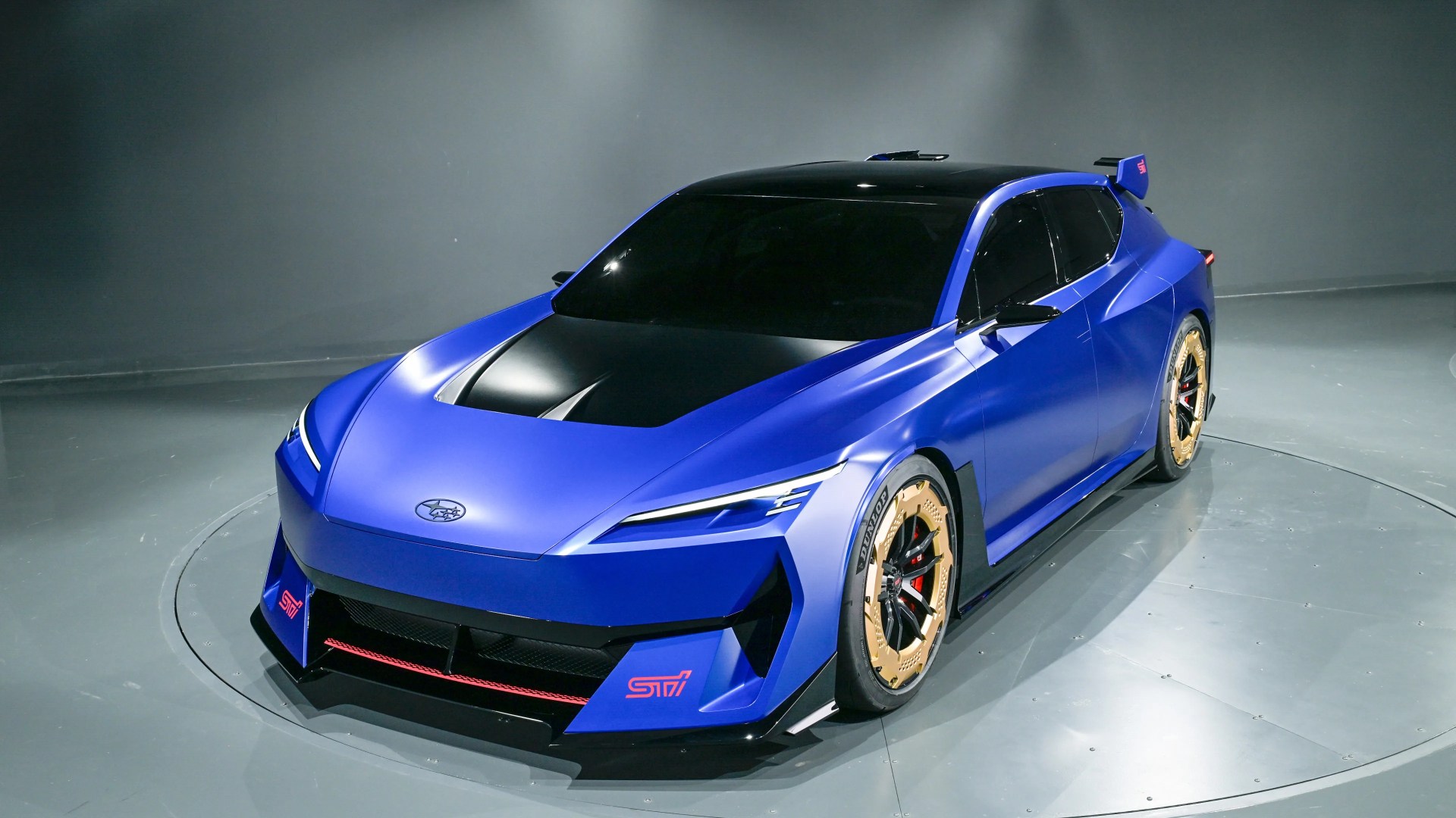 Matte blue Subaru sports car with black hood, gold wheels, and red STI logos in a studio setting.