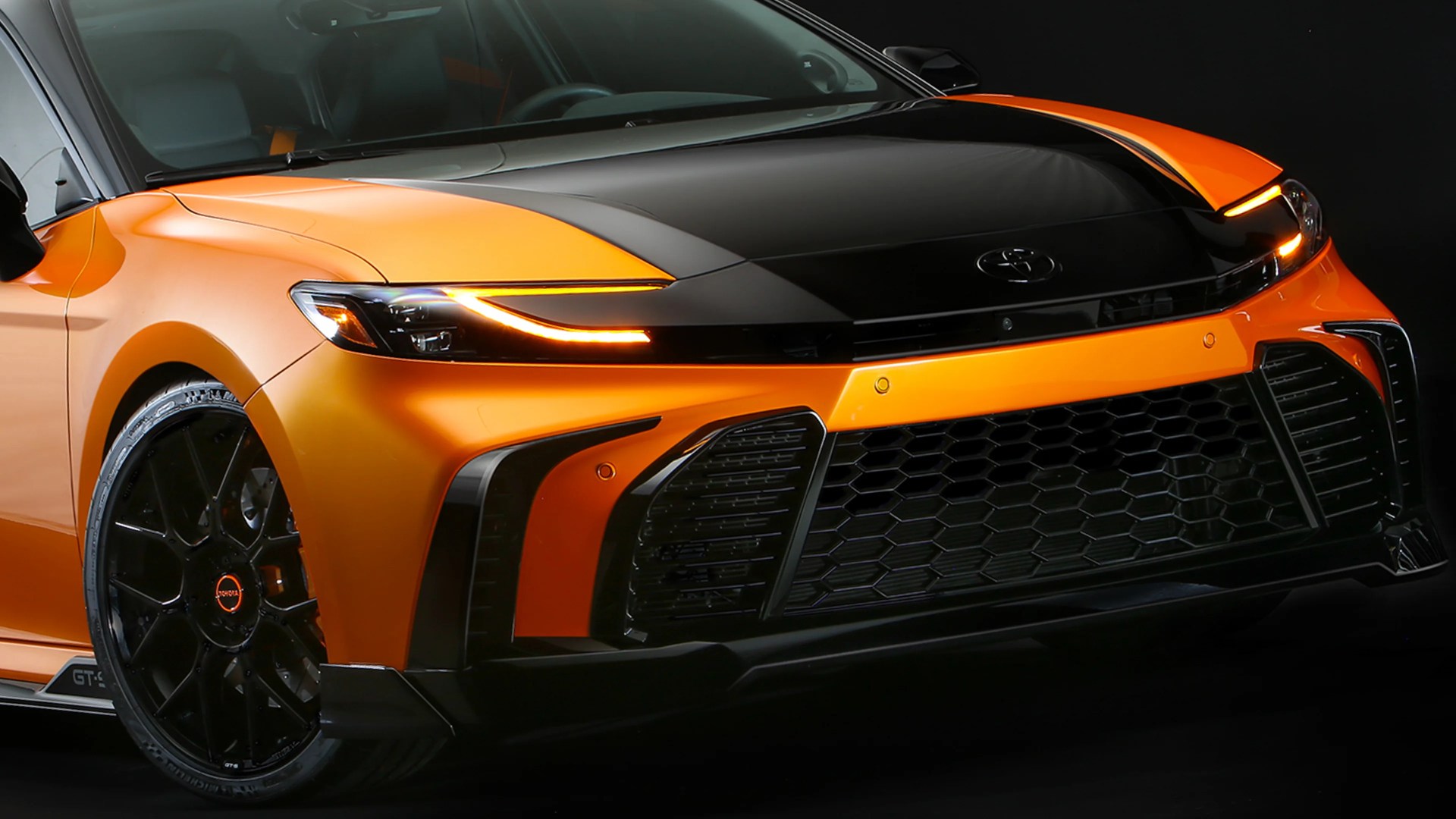 Front view of an orange and black Toyota sports car with illuminated LED headlights and black alloy wheels.