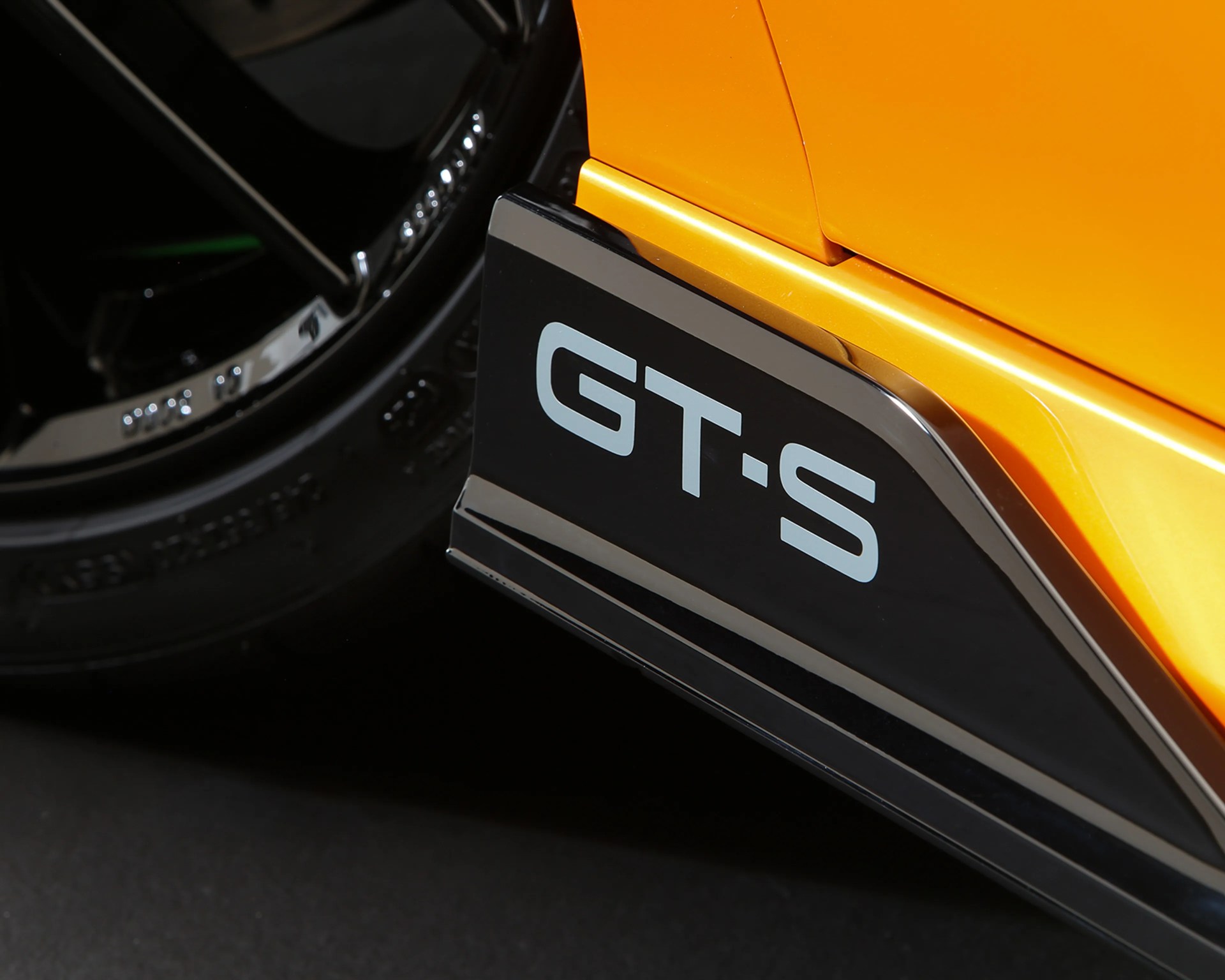 Close-up of a black side skirt with "GT·S" lettering on an orange car near a black wheel.