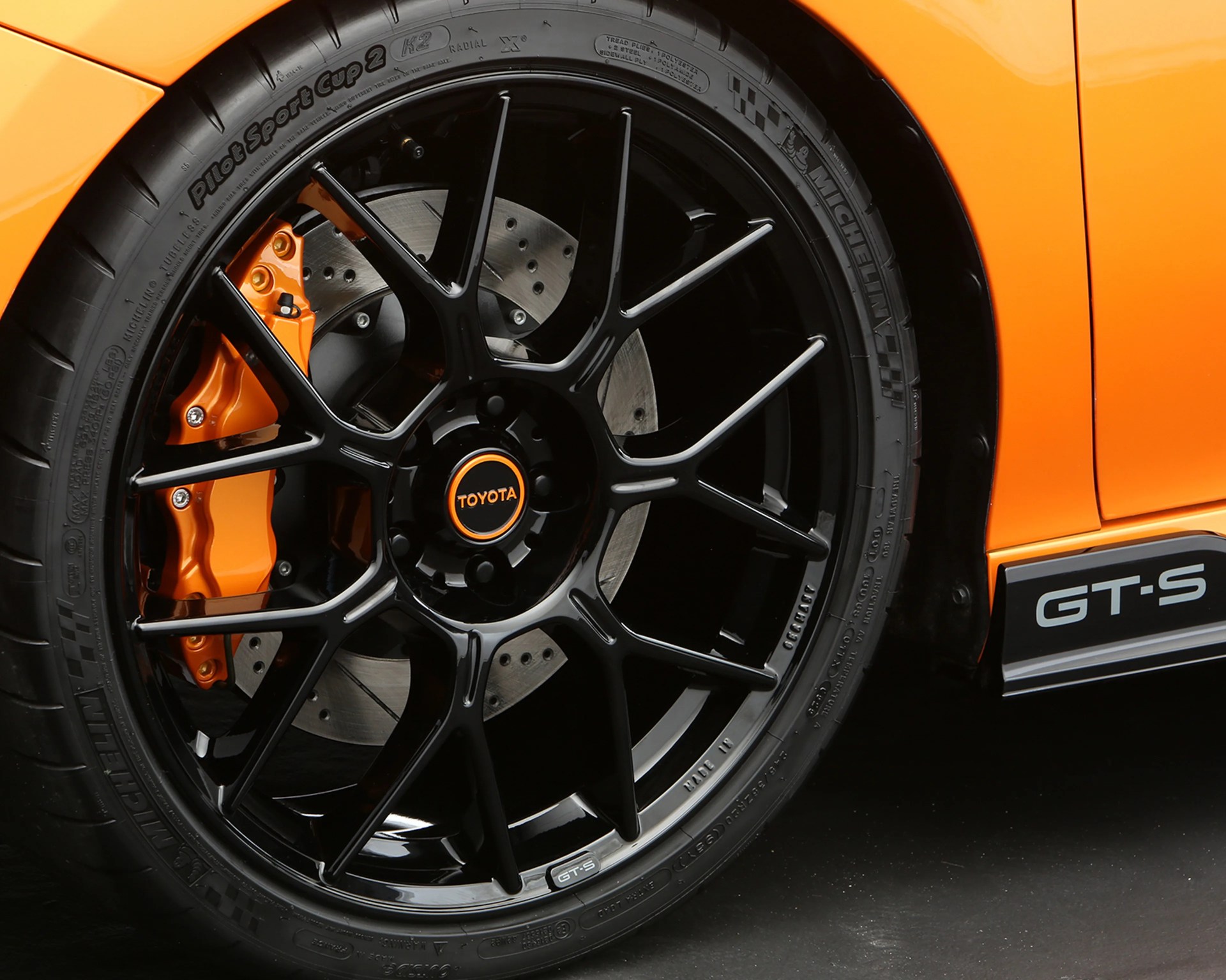 Glossy black multi-spoke Toyota wheel with orange brake caliper and Michelin Pilot Sport Cup 2 tire on an orange GT-S car.