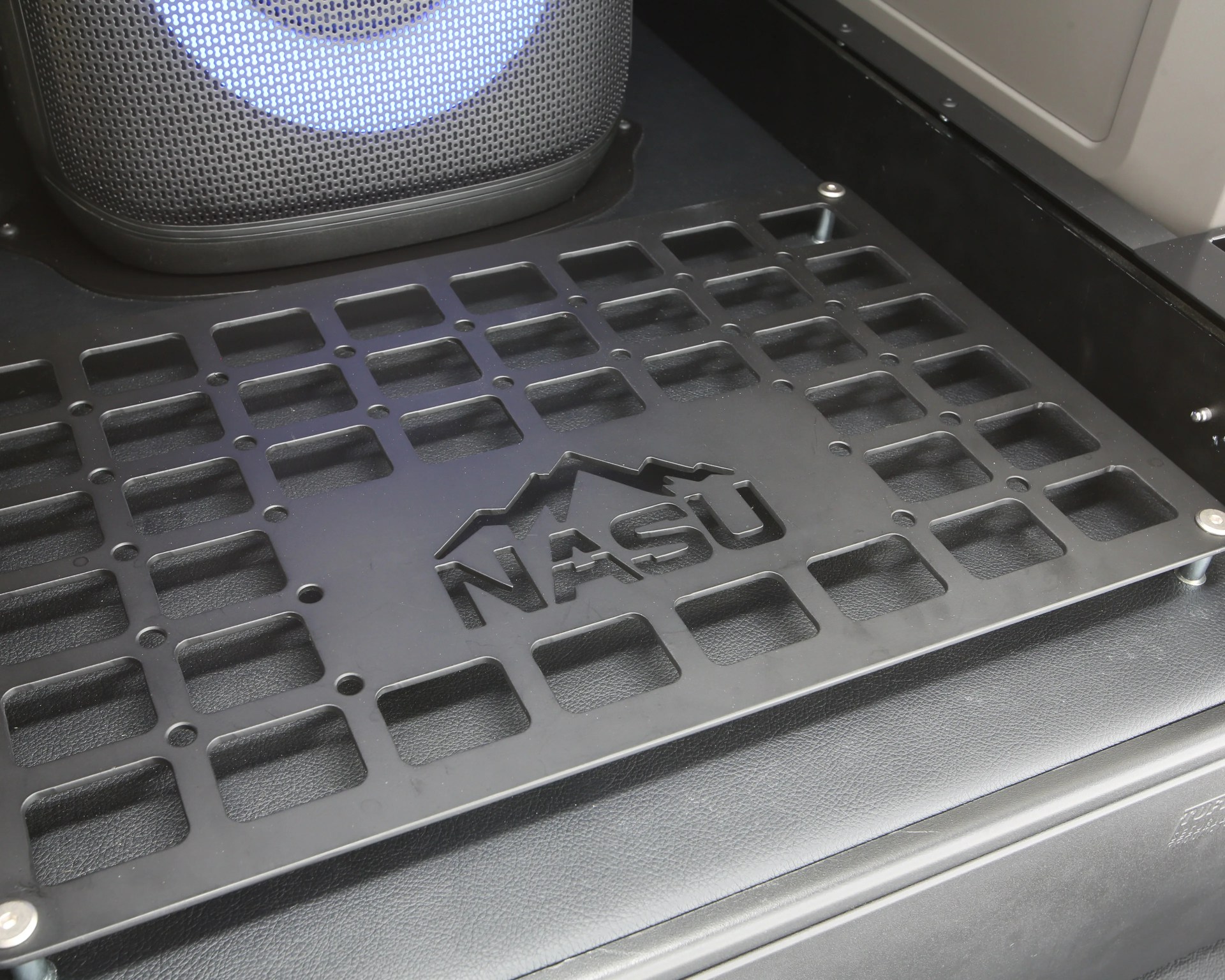 Black metal grid platform with the word "NASU" and mountain logo cut out, mounted on a textured black surface.