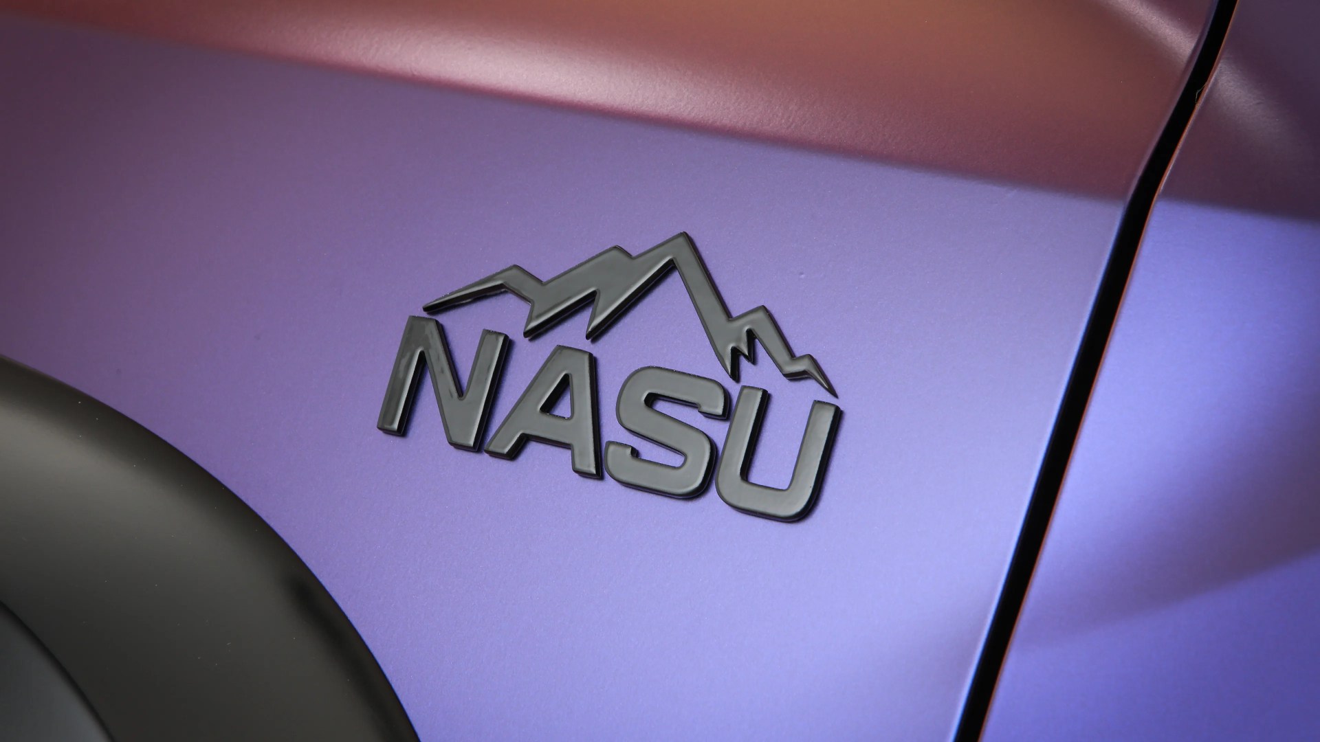 Glossy black "NASU" emblem with mountain design on a purple metallic car panel near a black fender.