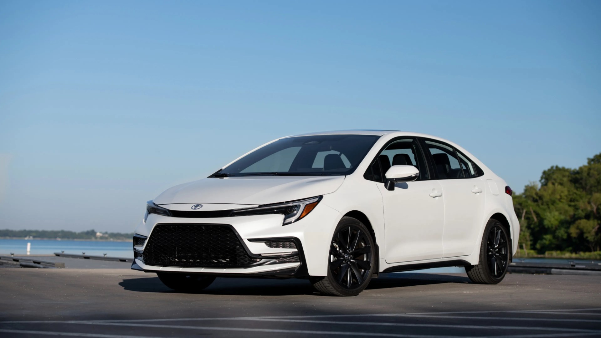White Toyota sedan with black wheels parked on a waterfront pavement under clear blue sky.