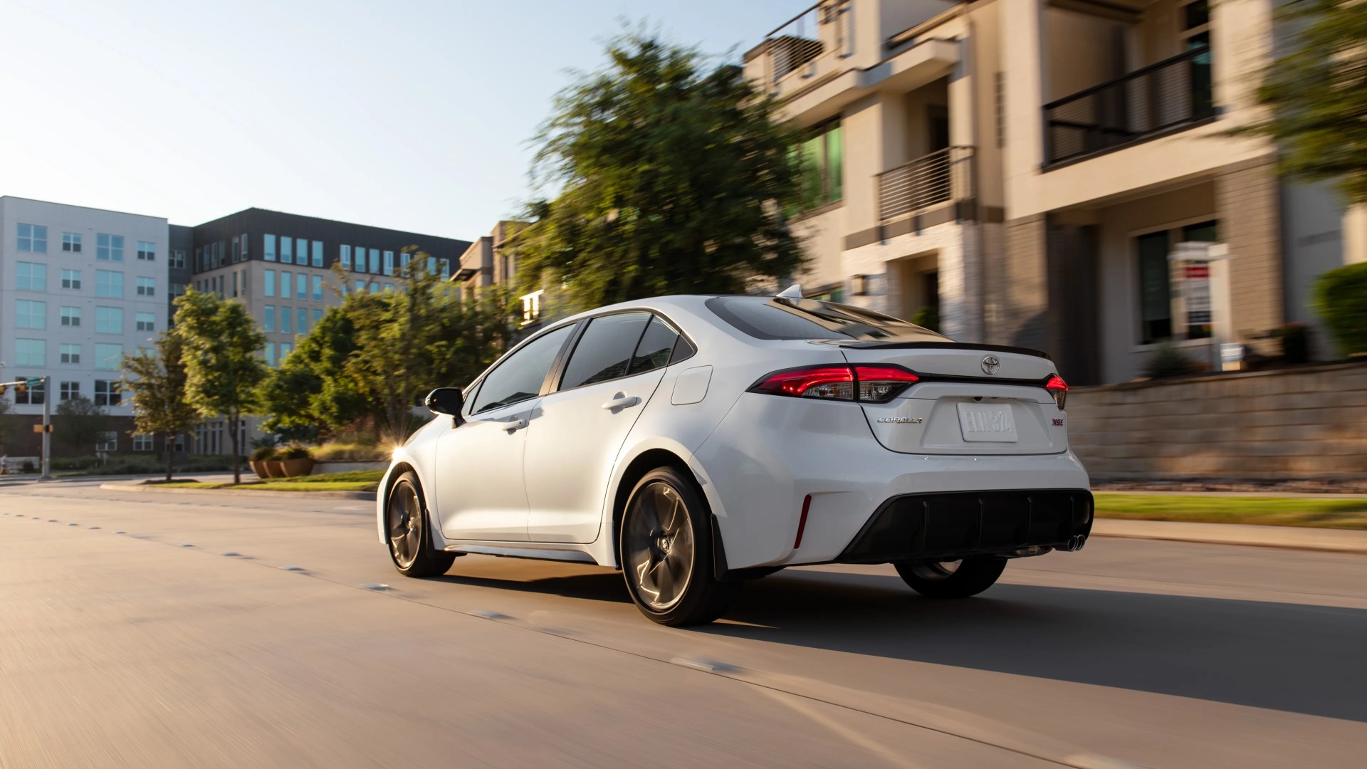 White Toyota Corolla XSE sedan driving on a city street with modern buildings and trees in the background.