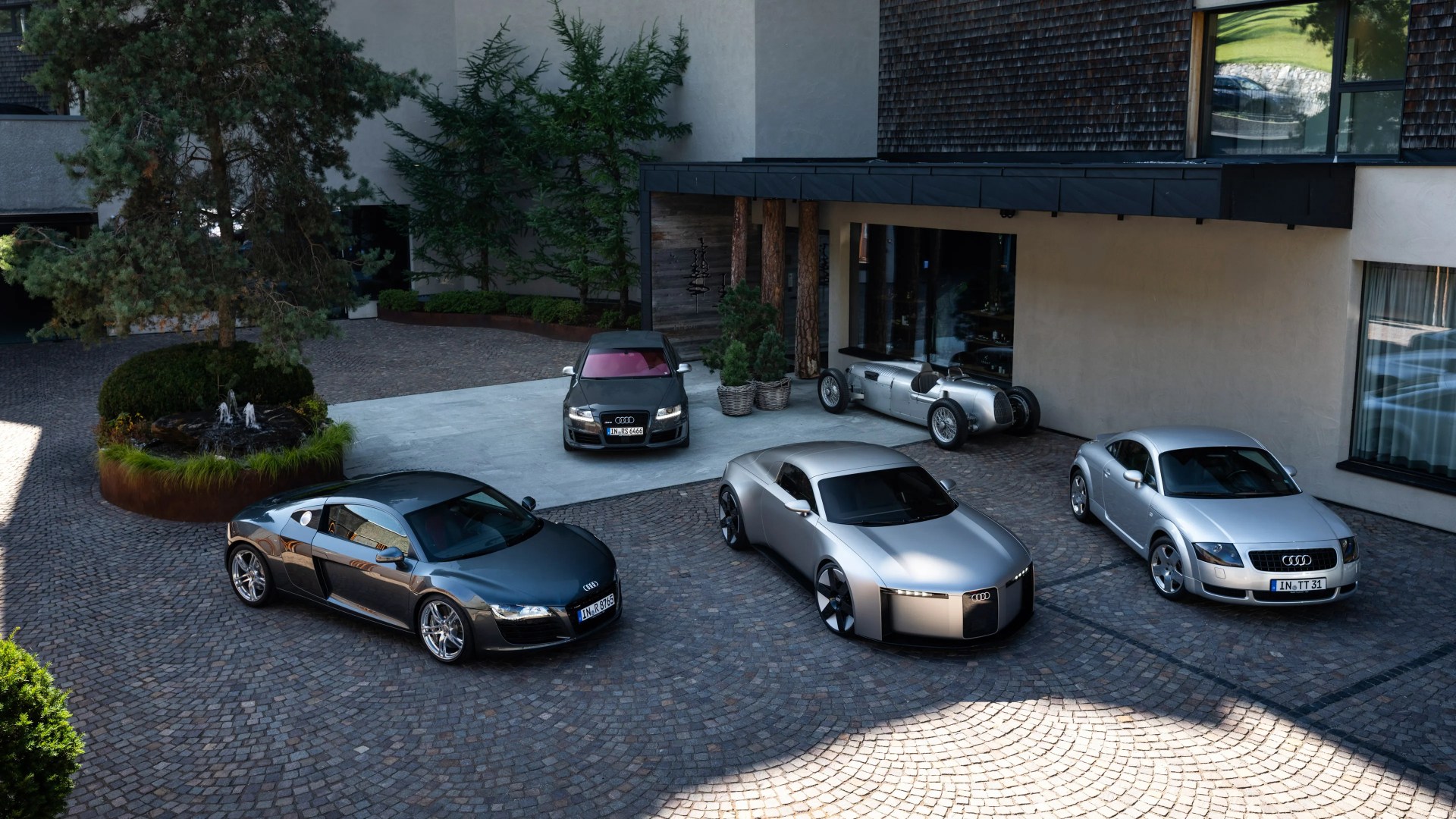 Five Audi cars parked on a cobblestone driveway in front of a modern building, including classic and futuristic models.