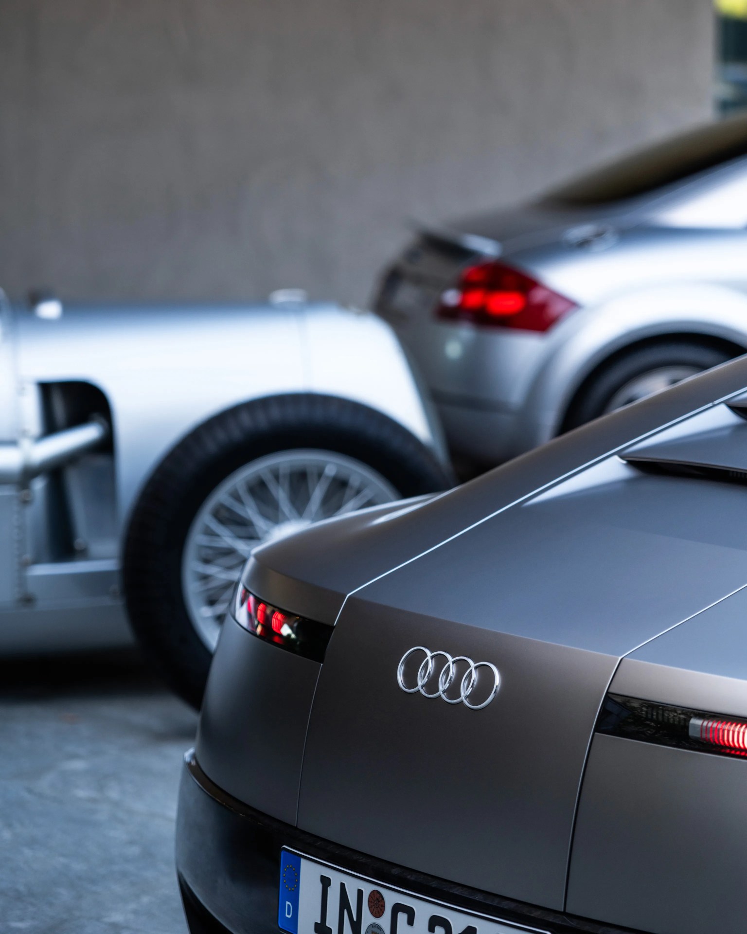Rear view of a matte gray Audi car with illuminated taillights and a European license plate, with two silver cars blurred in the background.