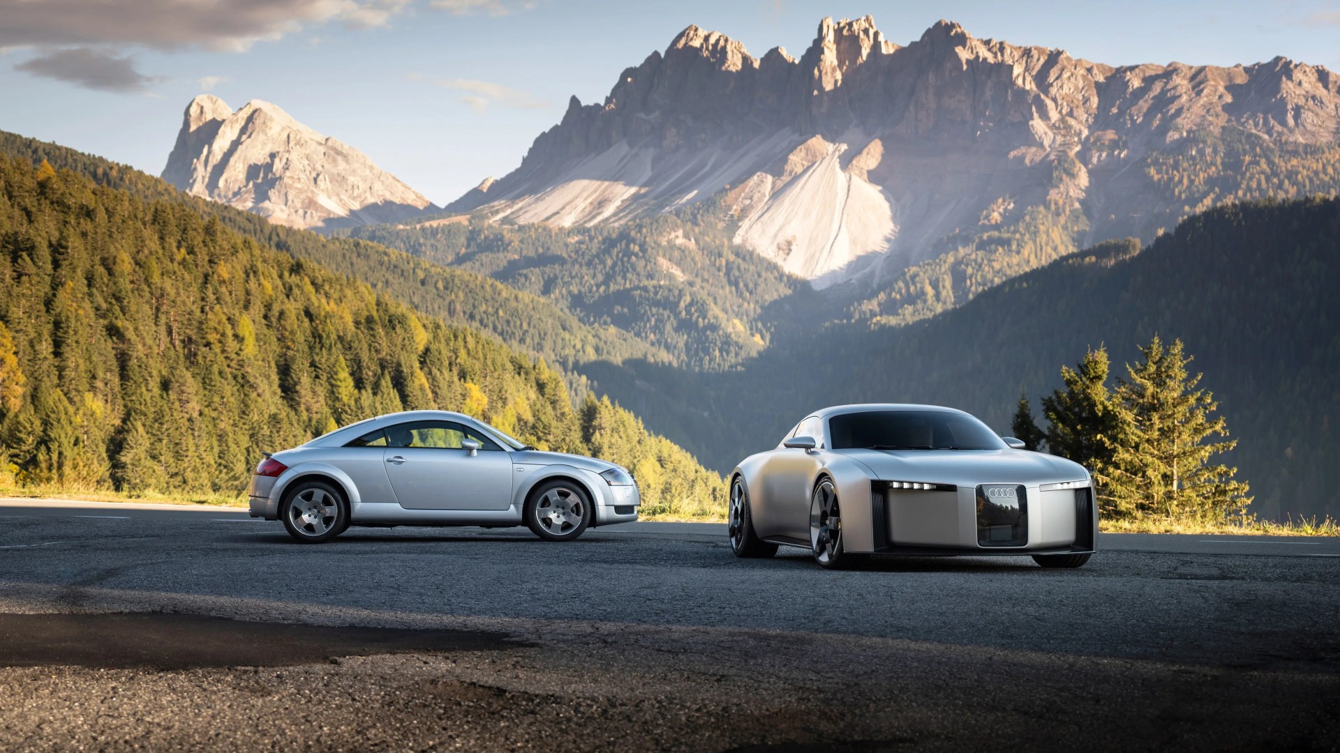 Two silver Audi sports cars, one classic and one modern concept, parked on a mountain road with forested hills and rocky peaks in the background.