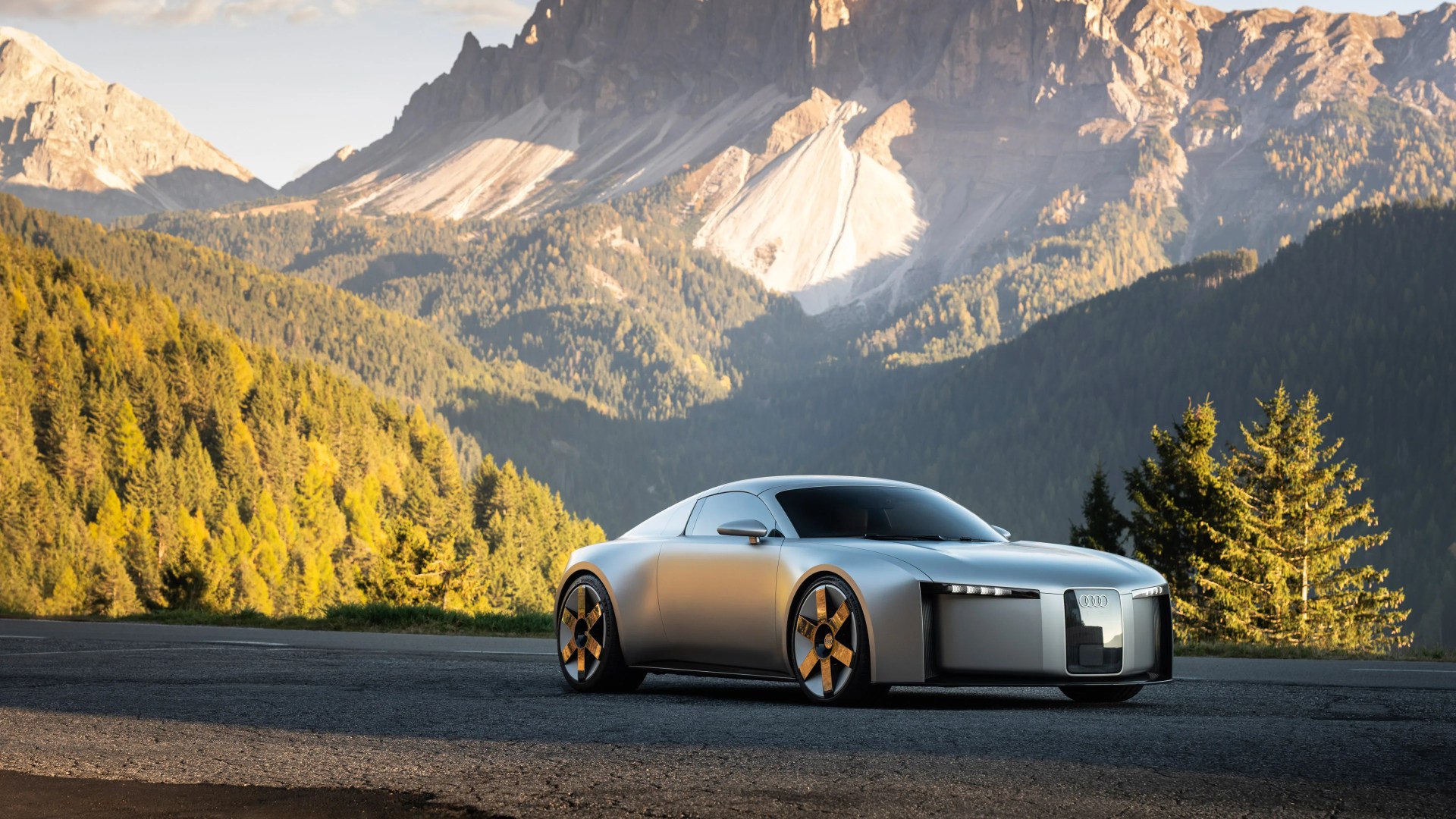 Silver Audi concept car with futuristic design parked on a mountain road with forested hills and rocky peaks in the background.