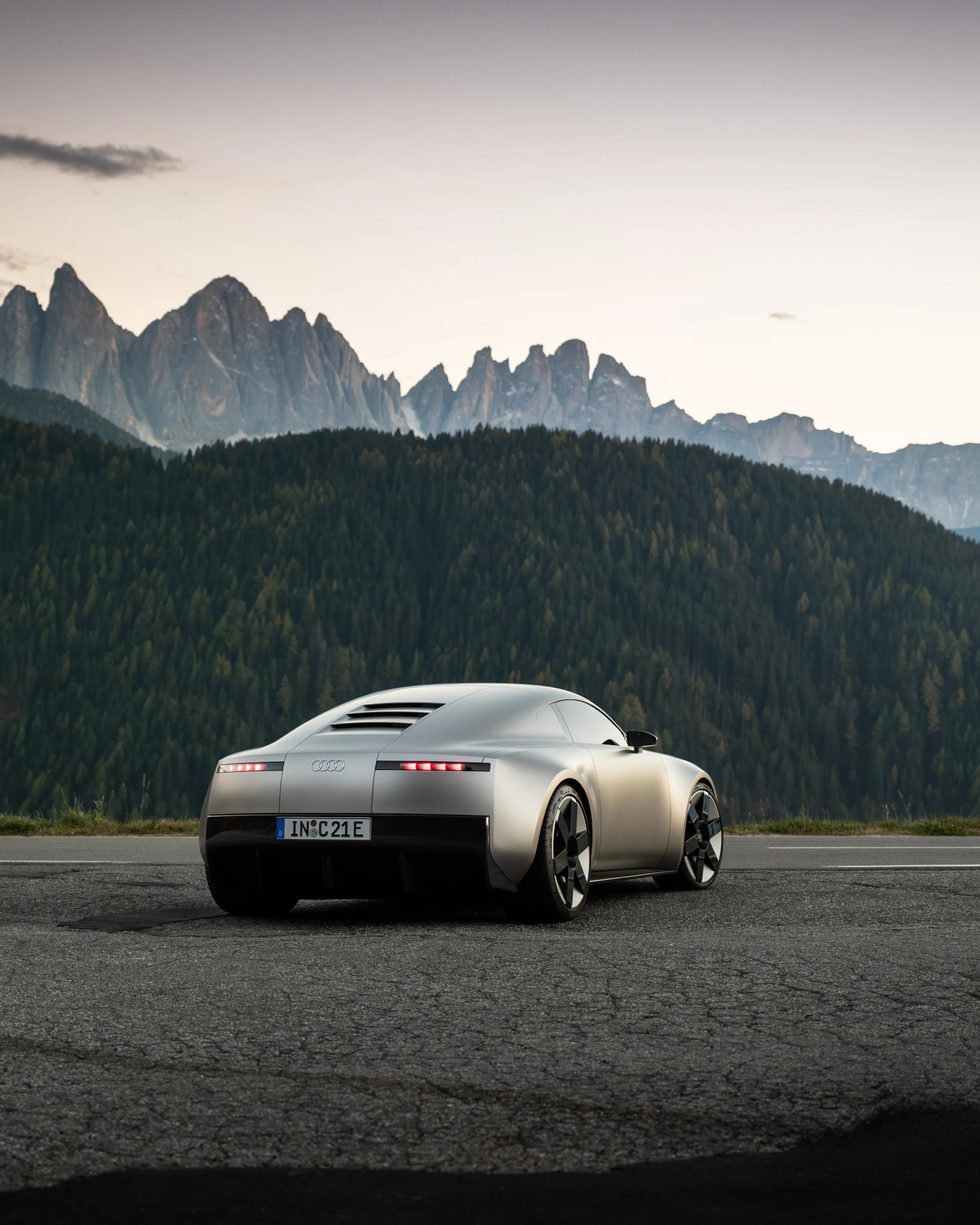 Silver Audi electric sports car parked on cracked asphalt with forested hills and mountain peaks in the background.