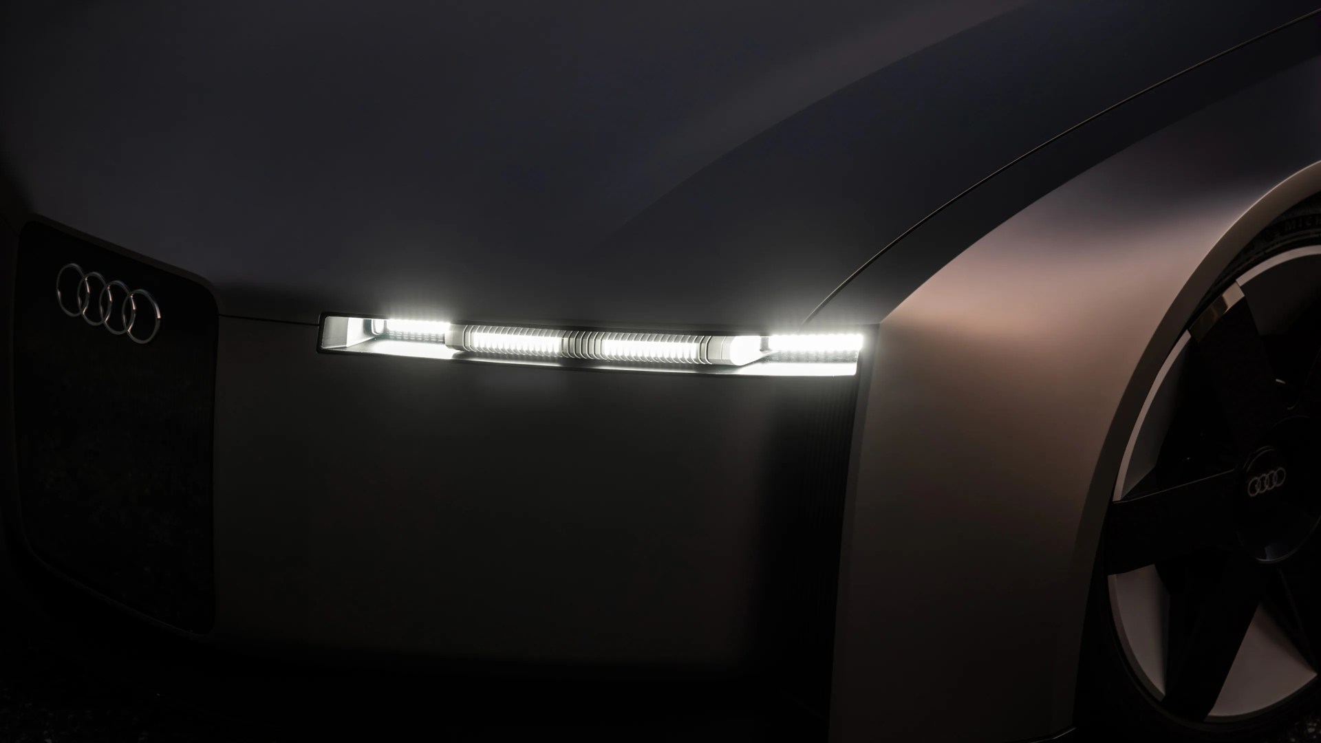 Close-up of a sleek gray Audi car front with illuminated horizontal LED headlight and black wheel with Audi logo.