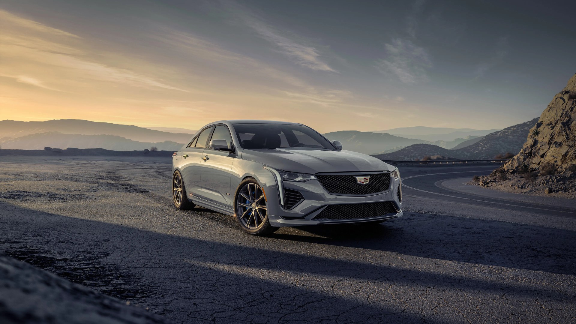 Gray Cadillac sedan parked on a cracked asphalt road with mountains and sunset in the background.