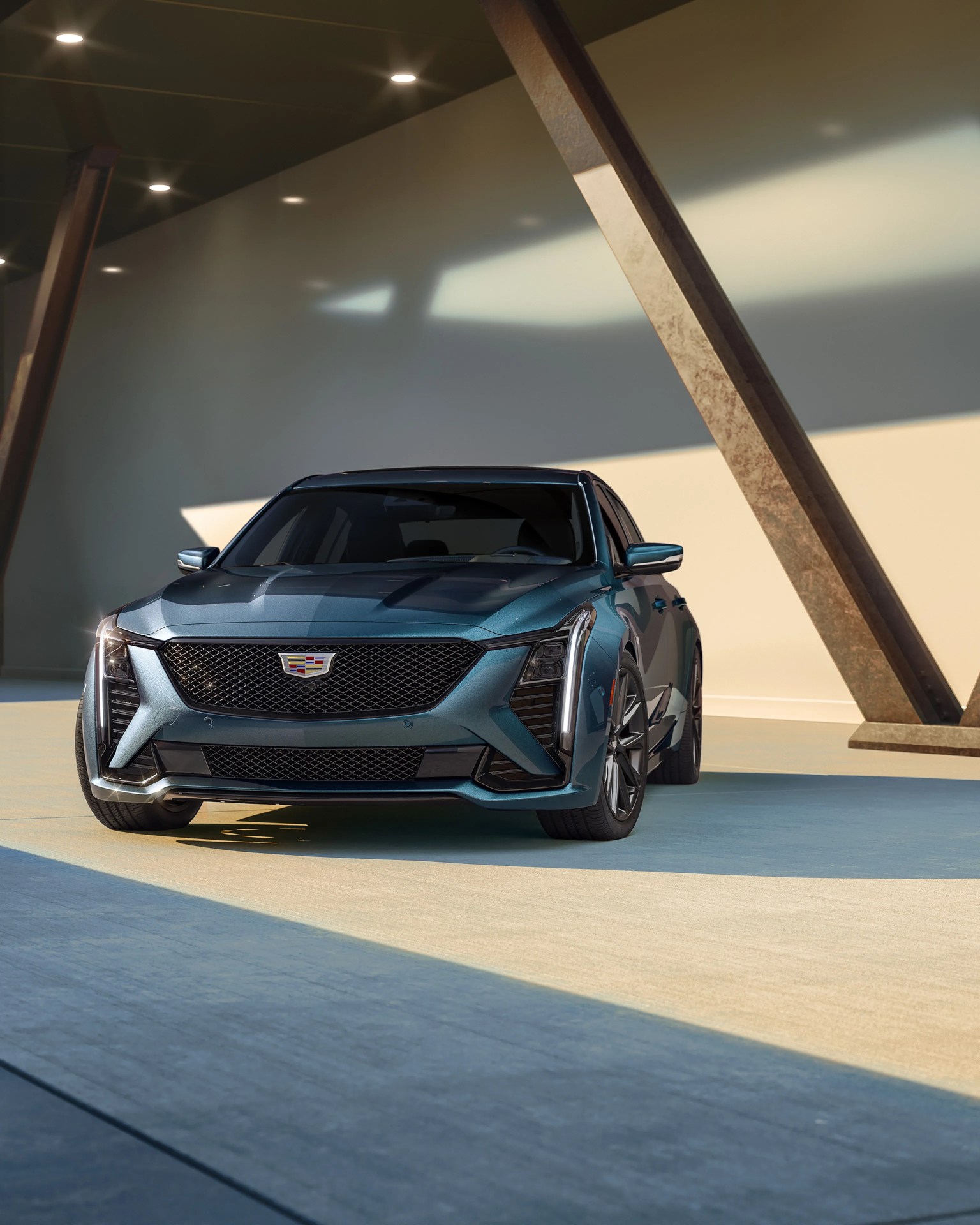 Metallic blue Cadillac sedan with black grille and vertical LED headlights in a modern, sunlit architectural setting.