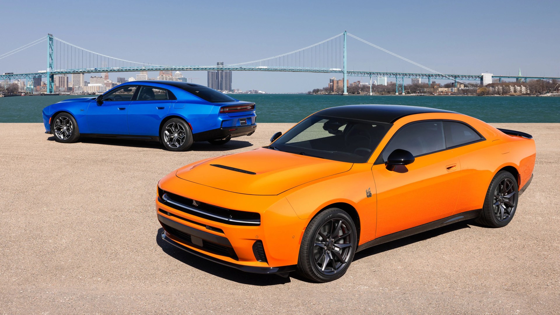 Bright orange Dodge Charger with black roof in front of a blue Dodge Charger parked near a river with a suspension bridge and city skyline in the background.