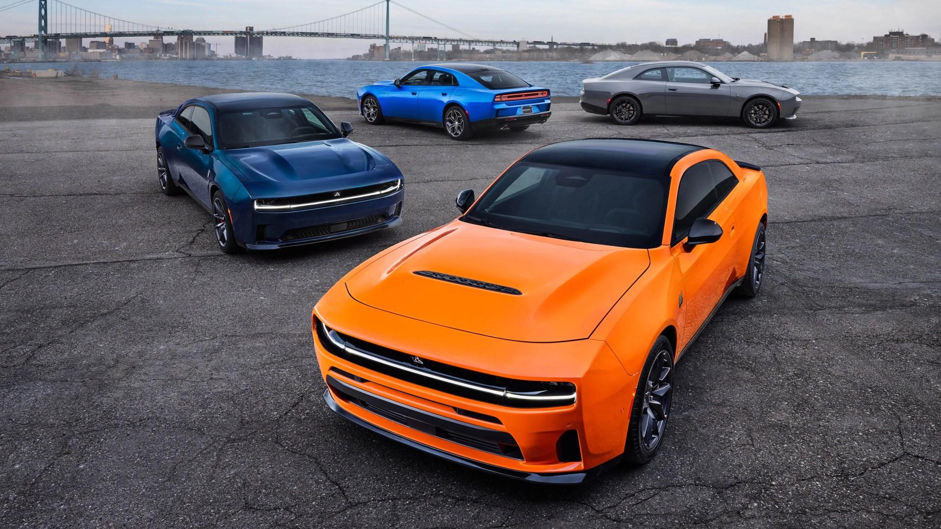 Four modern muscle cars in orange, blue, gray, and dark blue parked on cracked asphalt near a river with a city skyline and bridge in the background.