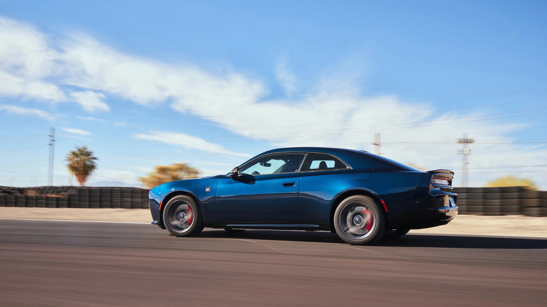 Dark blue Dodge Challenger coupe with red brake calipers driving on a racetrack under a blue sky.