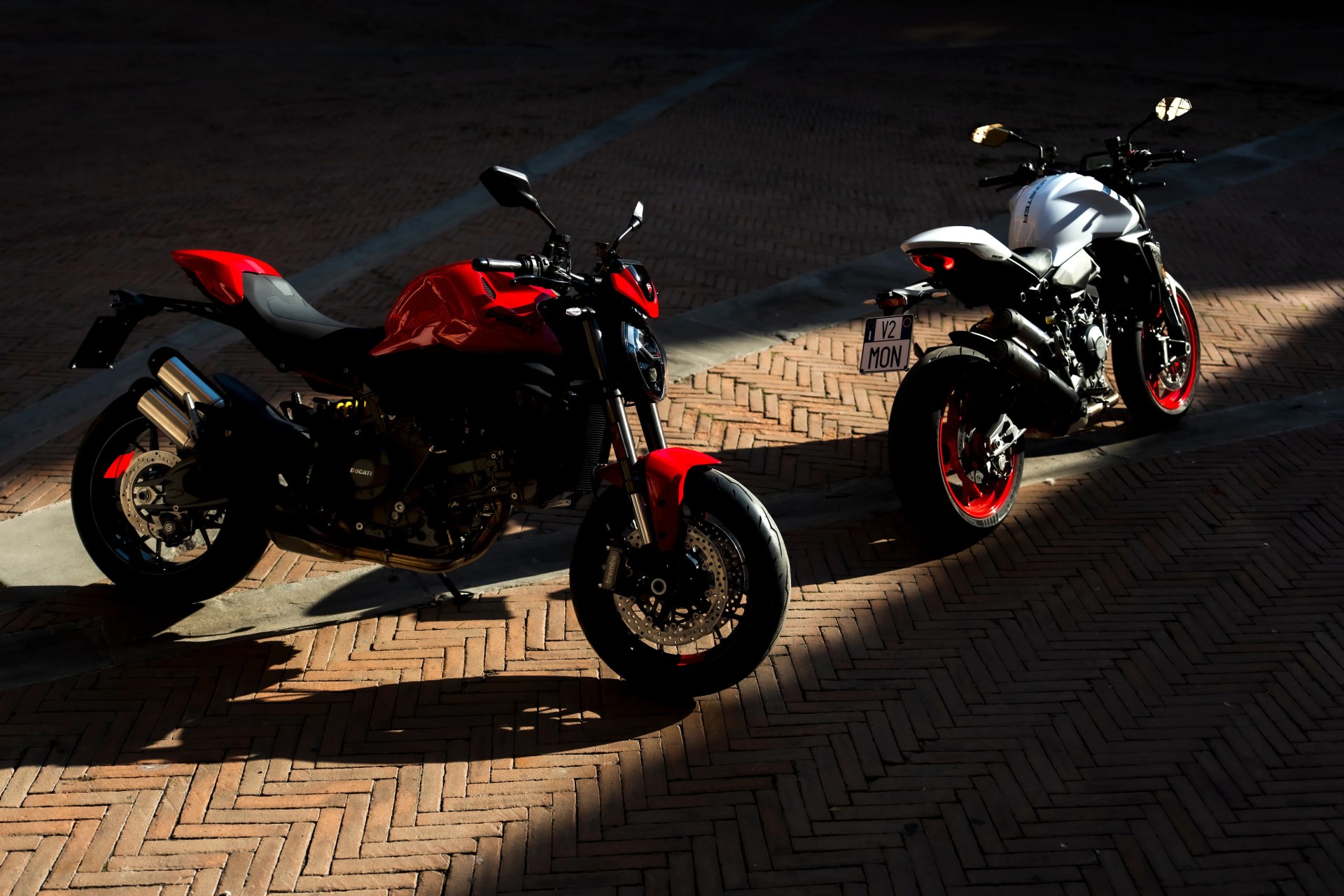 Two Ducati motorcycles, one red and one white with red rims, parked on a brick pavement in sunlight and shadow.