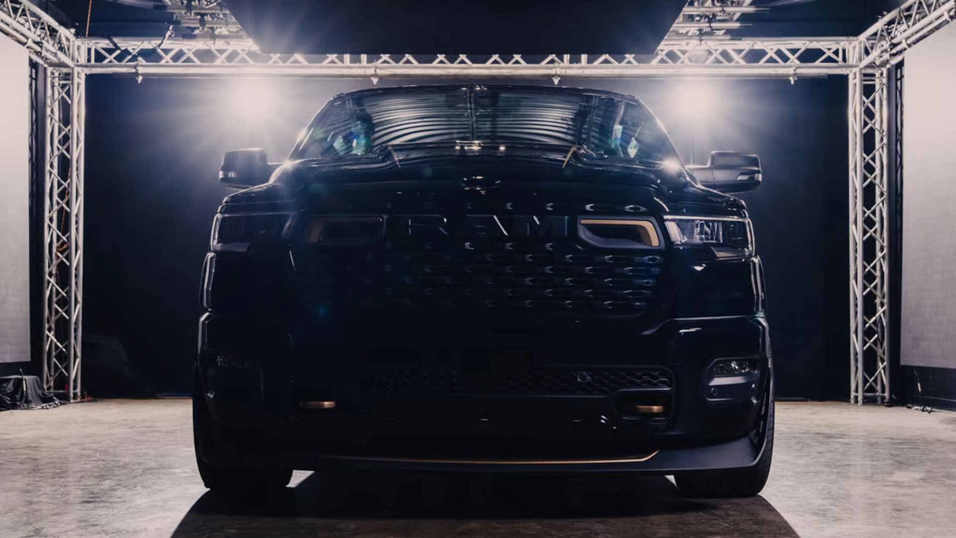 Front view of a black Ram pickup truck under bright studio lights with a metal truss frame background.