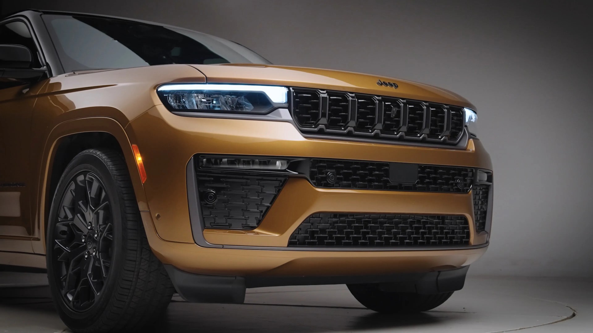 Front view of a gold Jeep SUV with LED headlights and black grille in a studio setting.