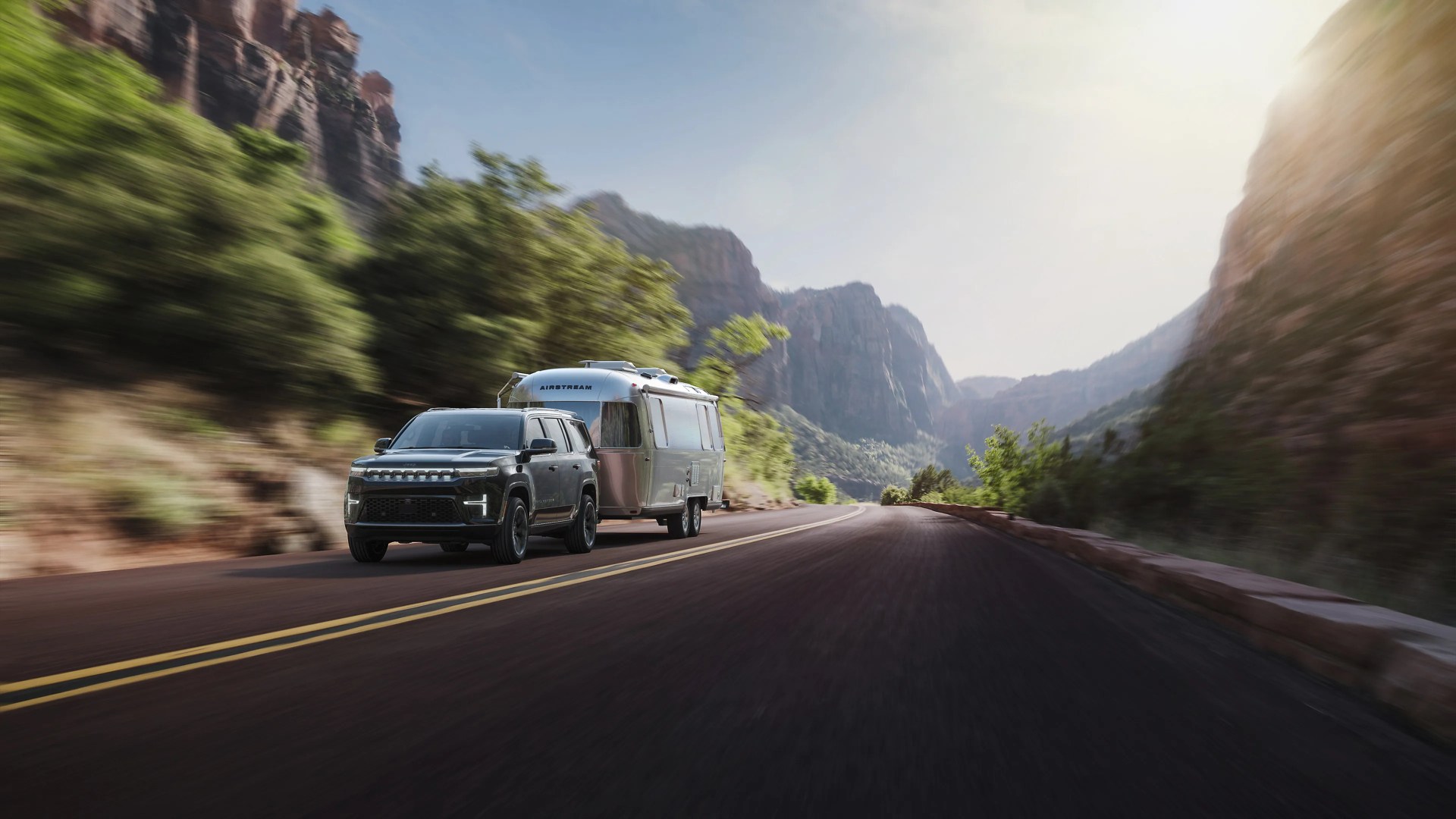 Black SUV towing a silver Airstream trailer on a mountain road with cliffs and trees.