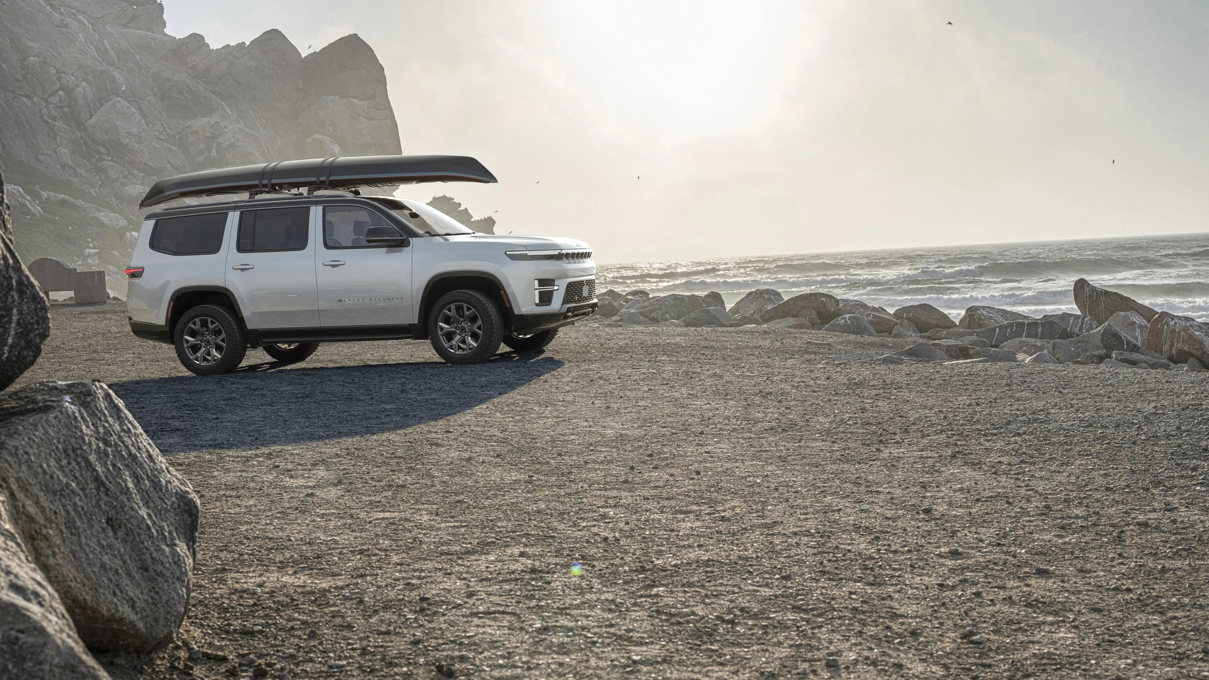 White SUV with a canoe mounted on the roof parked on a rocky beach near the ocean.