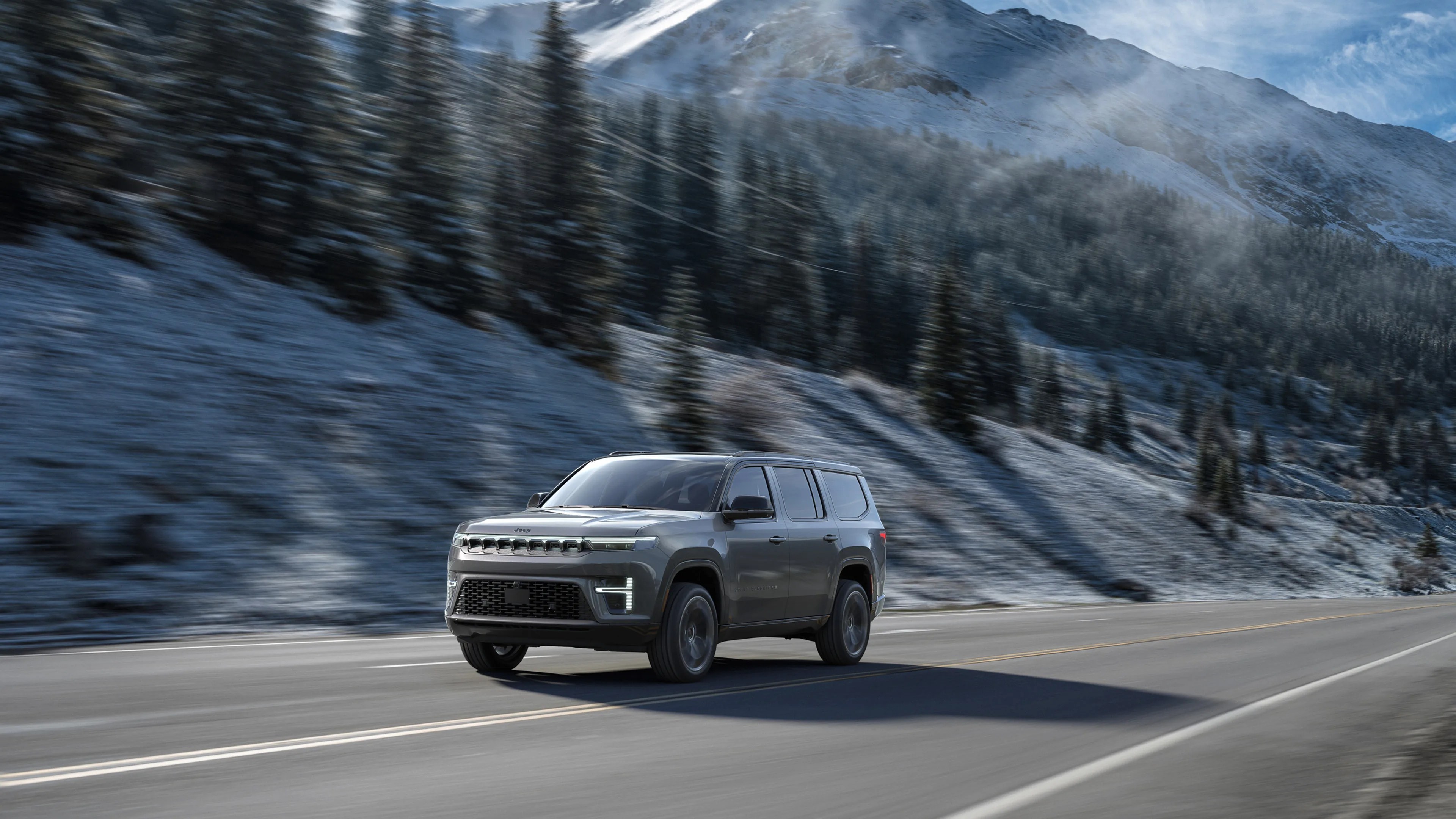 Gray Jeep SUV driving on a mountain road with snow-covered trees and peaks in the background.