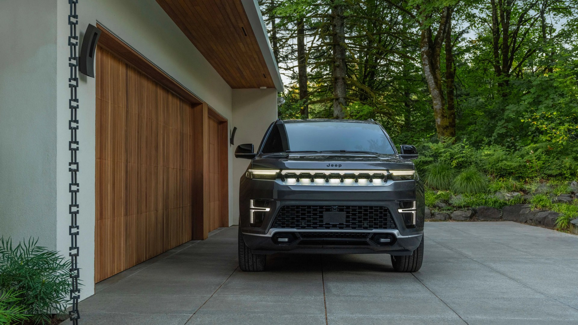Black Jeep SUV with illuminated front grille and headlights parked in a driveway next to a wooden garage door.