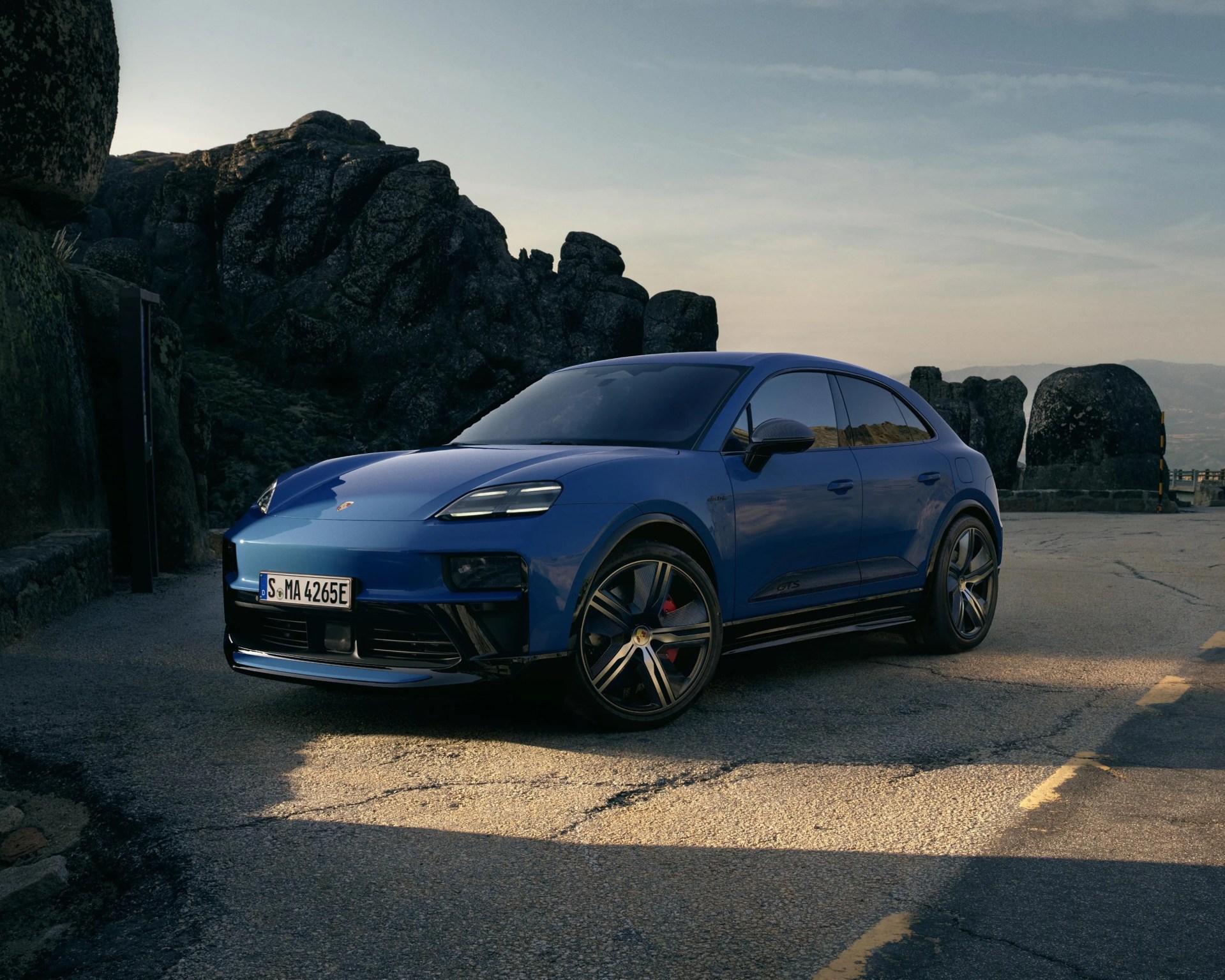 Blue Porsche electric SUV parked on a mountain road with rocky cliffs in the background.