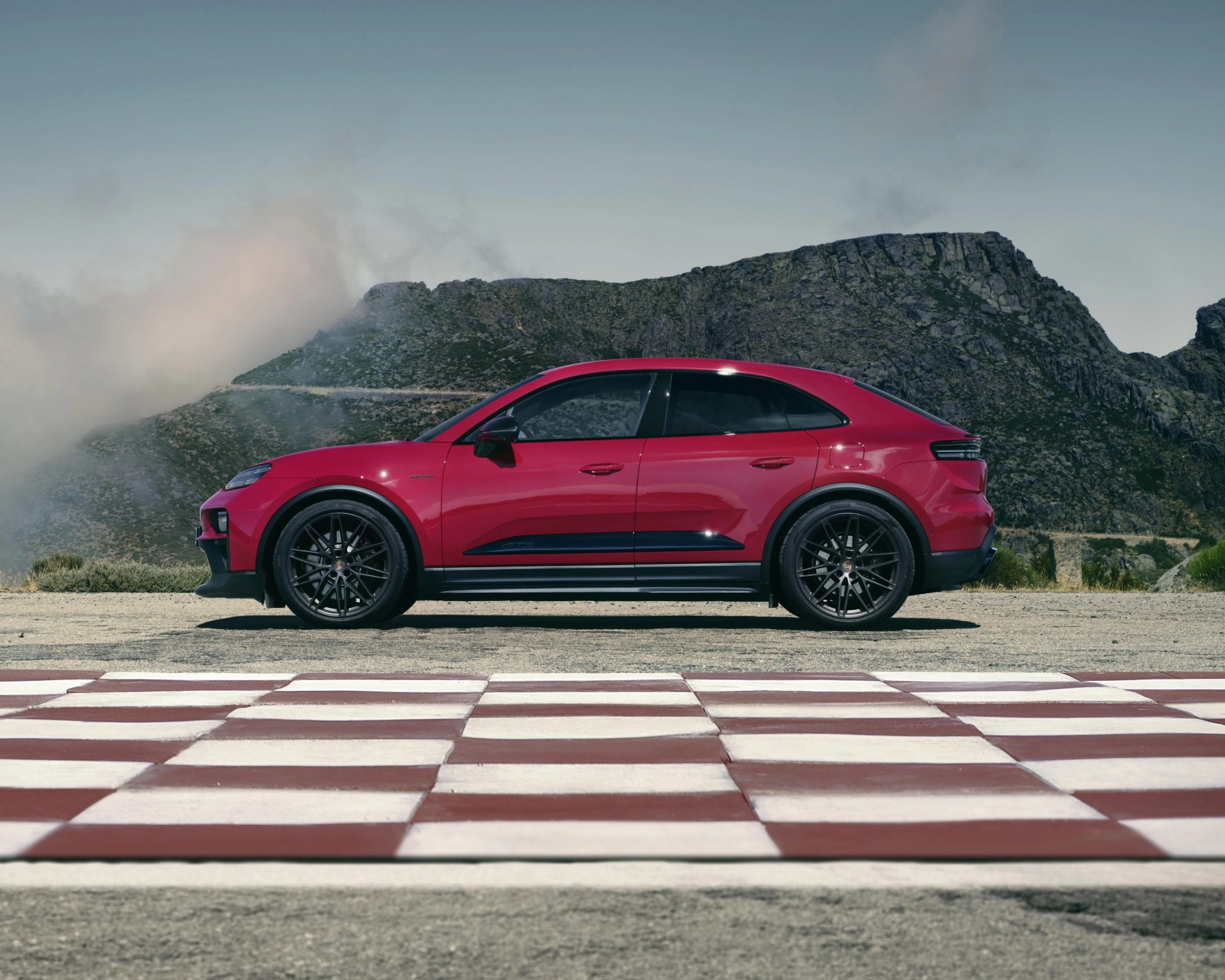 Red sporty SUV with black wheels parked on a checkered surface with rocky hills in the background.