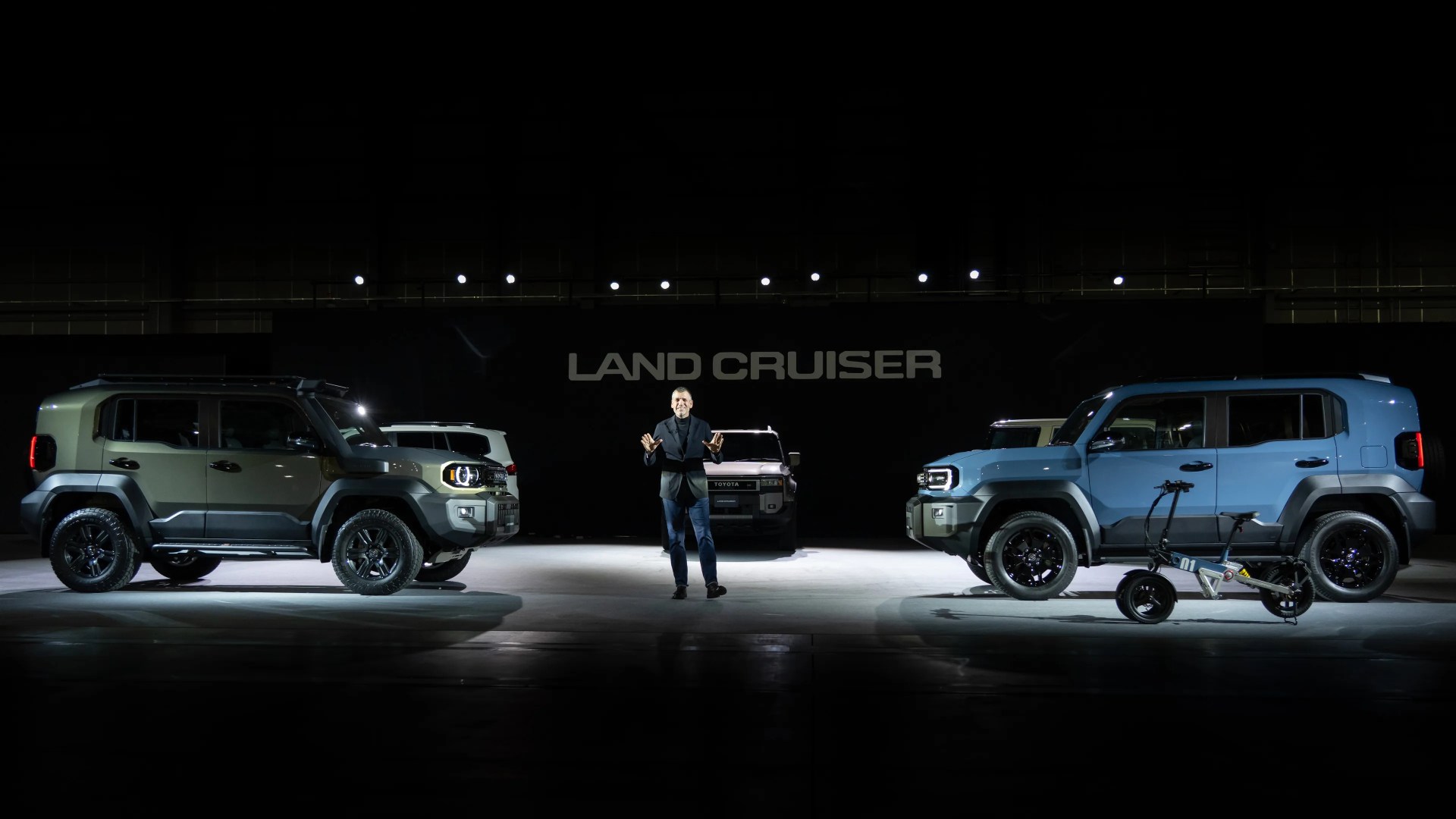 Man standing between two Toyota Land Cruiser SUVs, one green and one blue, with a small electric scooter in front of the blue SUV.