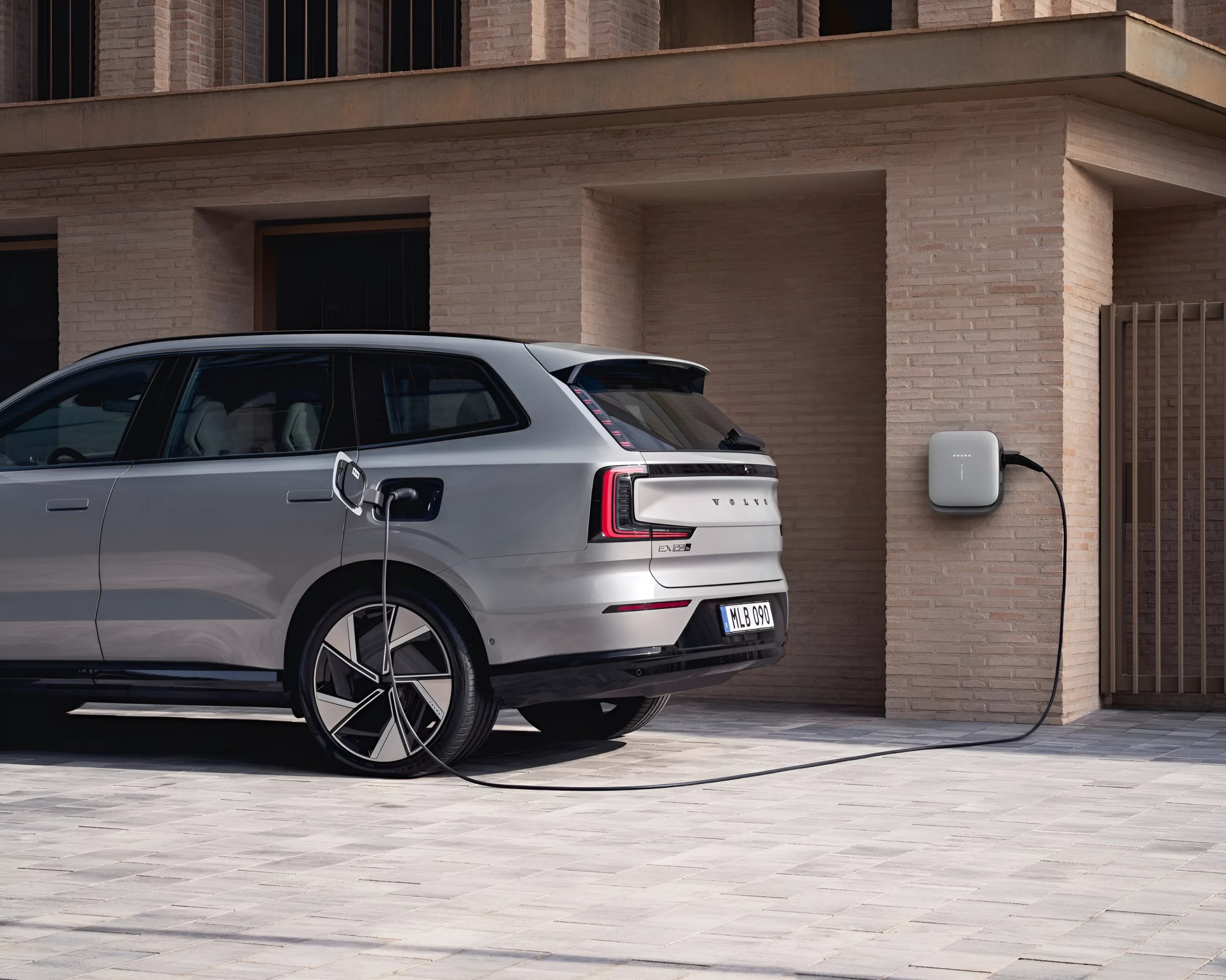 Silver Volvo EX90 electric SUV charging at a wall-mounted charger outside a modern brick building.