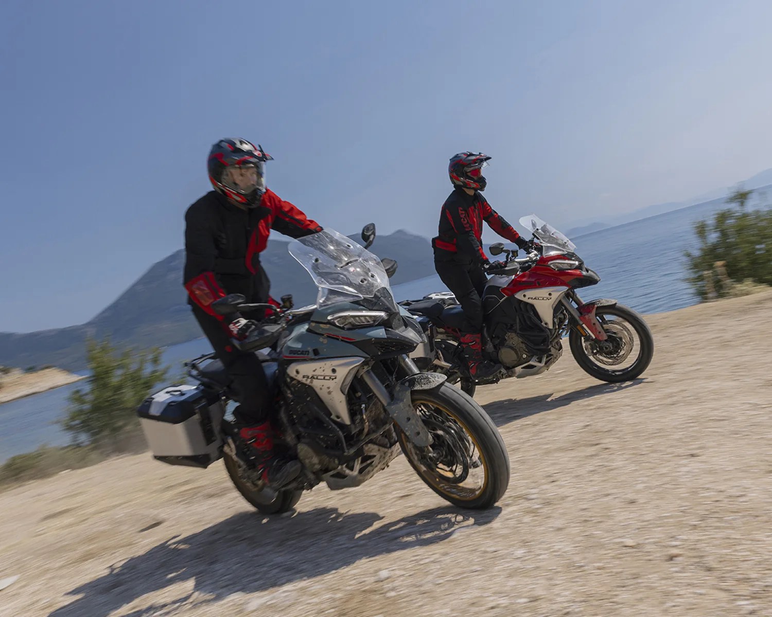 Two riders in black and red gear on Ducati adventure motorcycles riding on a dirt path near a body of water.