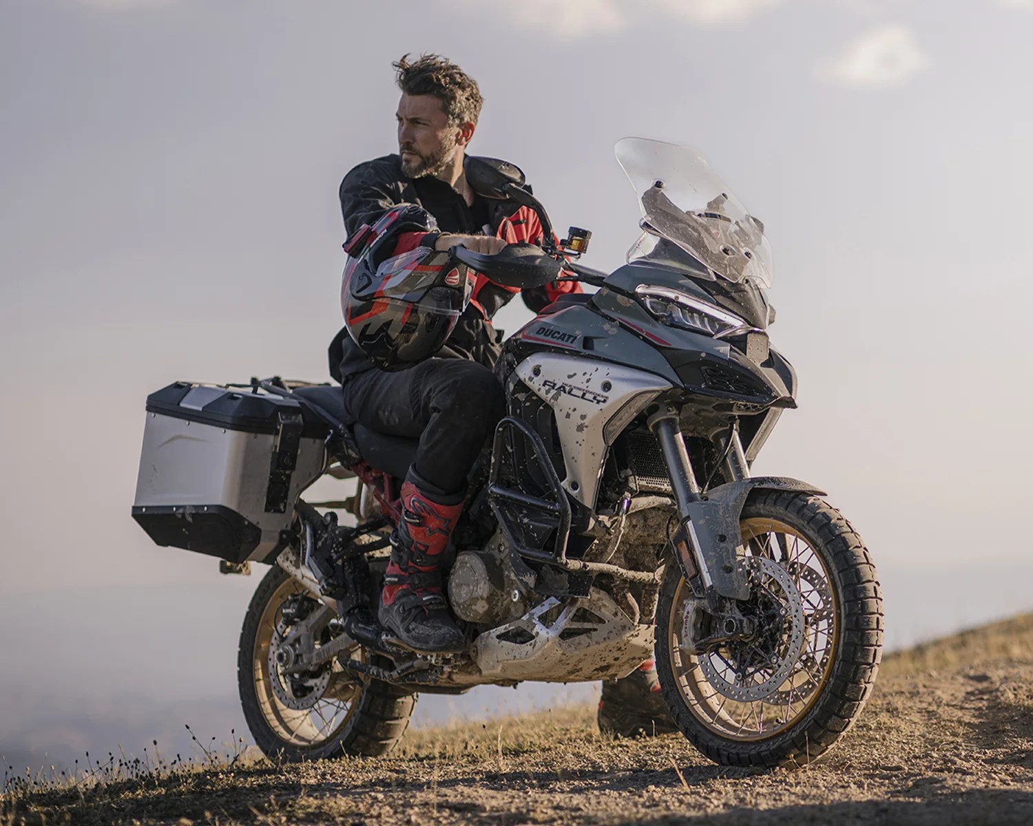 Man in black and red riding gear sitting on a muddy silver and black Ducati Rally motorcycle on a dirt path.