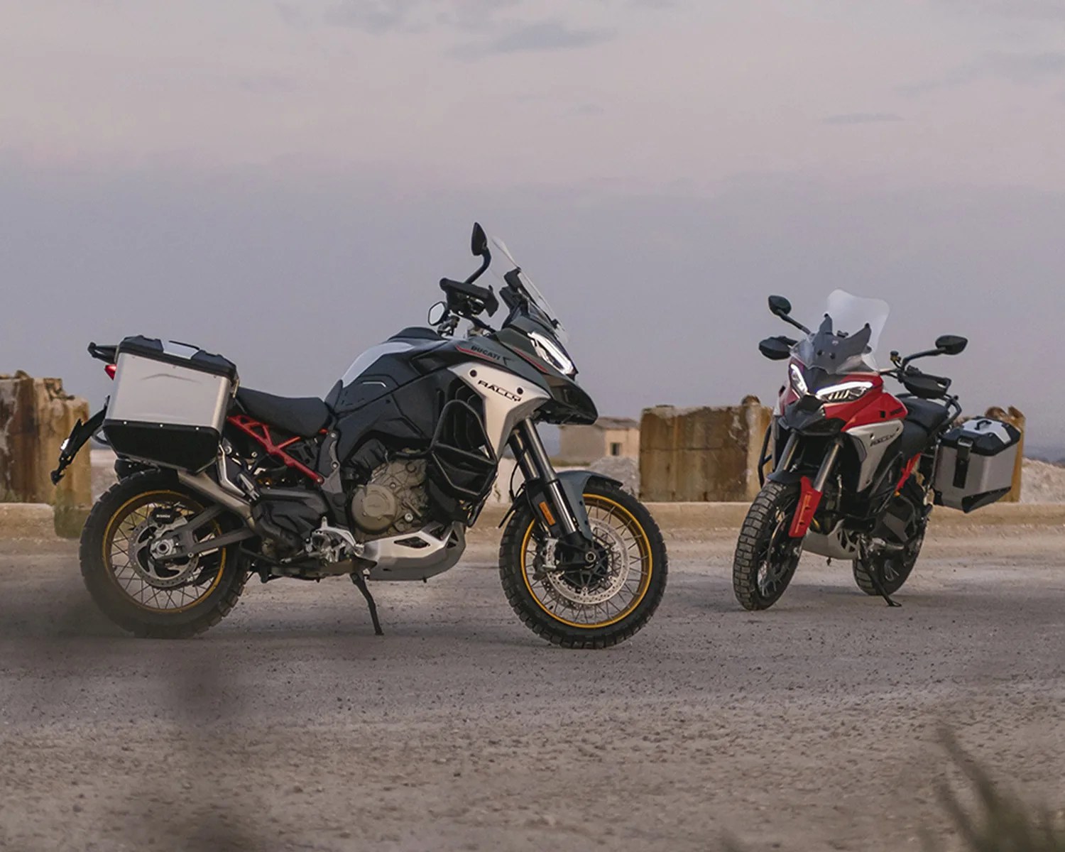 Two adventure motorcycles with side panniers, one black and white with gold rims, the other red and black, parked on a gravel surface.