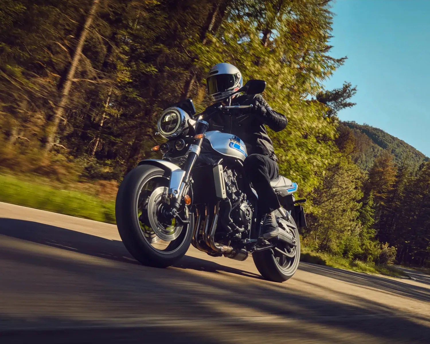 Motorcyclist wearing black gear and silver helmet riding a silver and blue Honda motorcycle on a forested road.