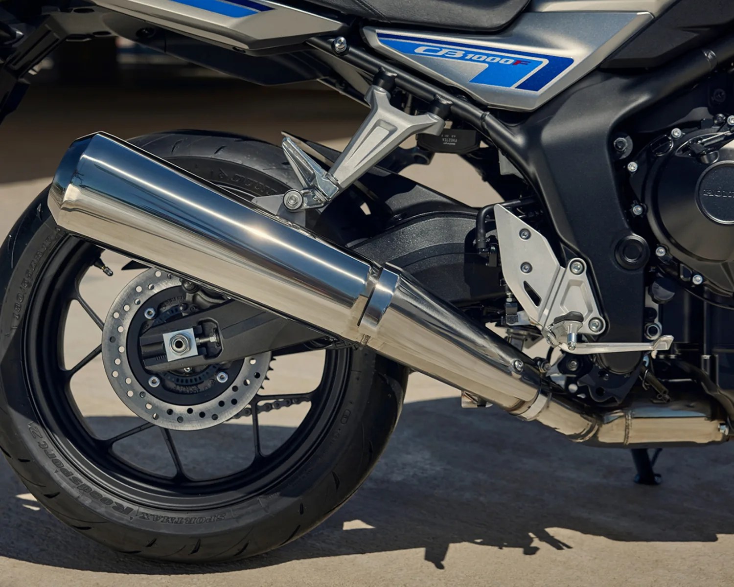 Close-up of a silver exhaust pipe and rear wheel of a blue and silver motorcycle with "CB1000F" decal.