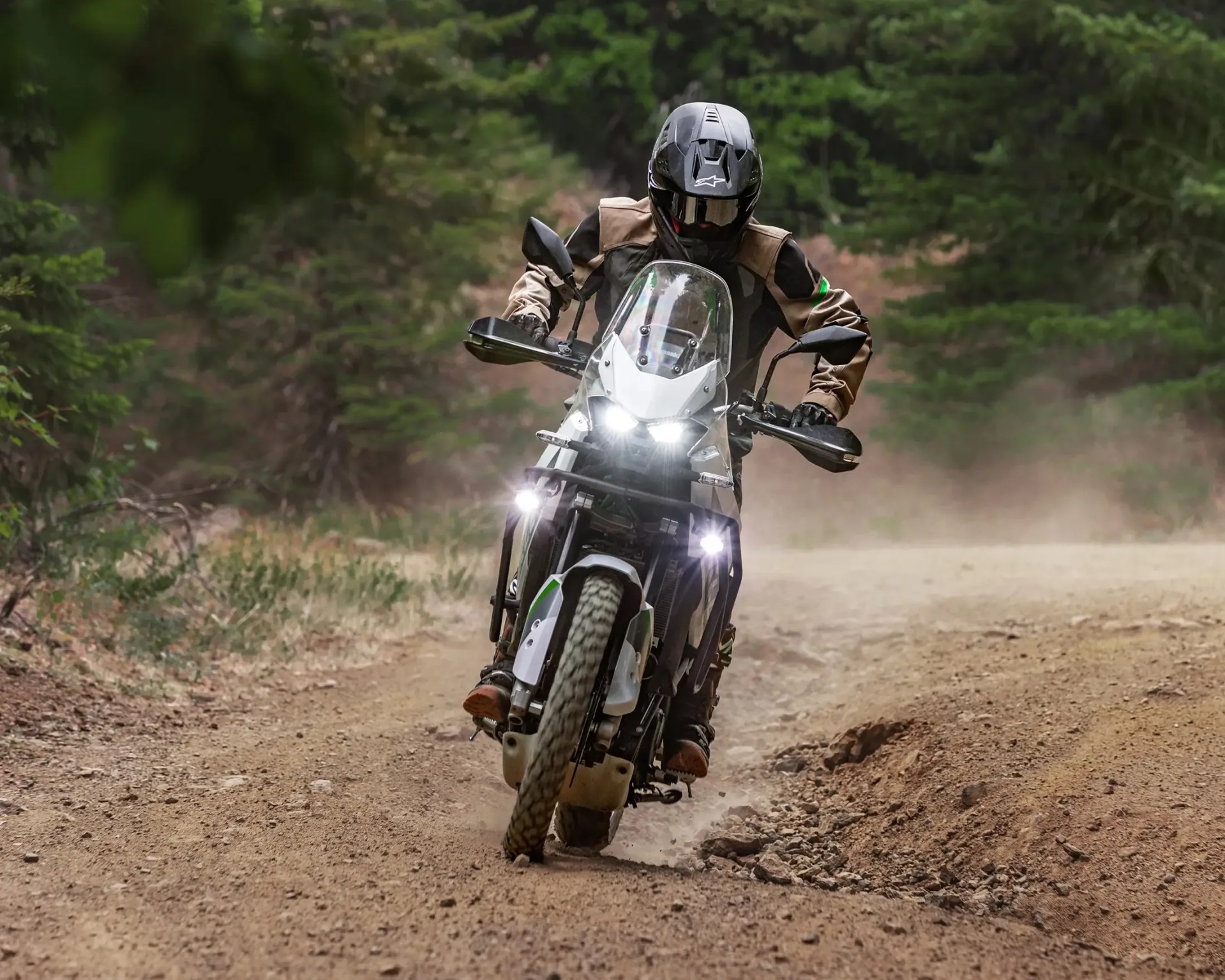 Motorcyclist wearing black helmet and tan jacket riding a white adventure bike on a dirt trail in a forest.