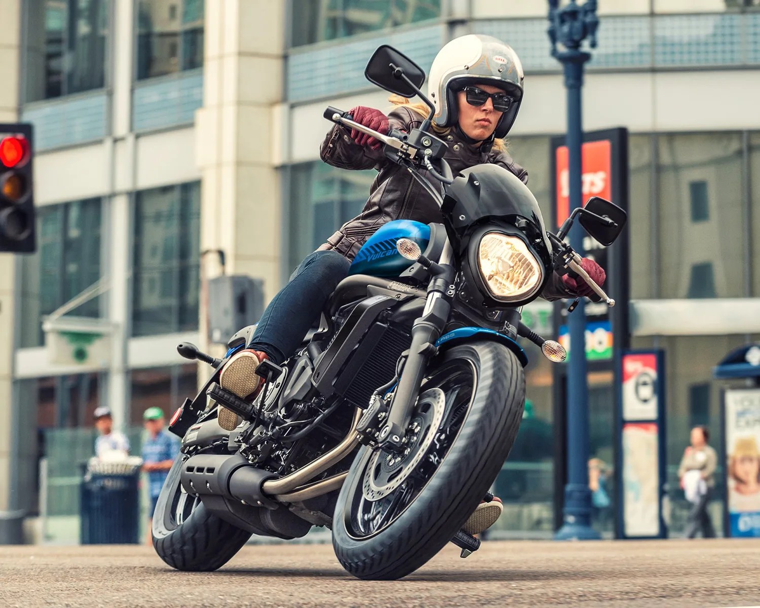Person wearing a white helmet and brown jacket riding a blue and black Vulcan motorcycle on a city street.