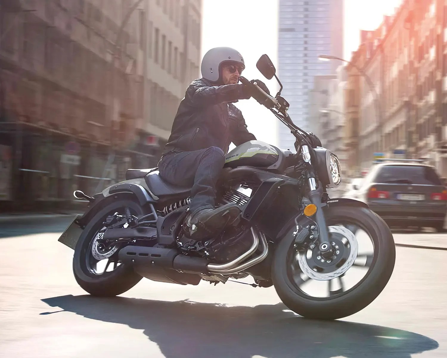 Man wearing a gray helmet and black jacket riding a black and gray motorcycle on a city street.