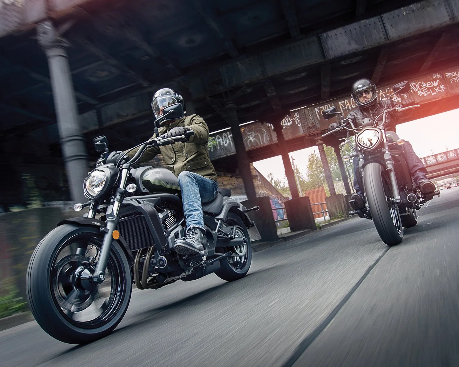 Two riders on black and gray cruiser motorcycles under a graffiti-covered bridge.