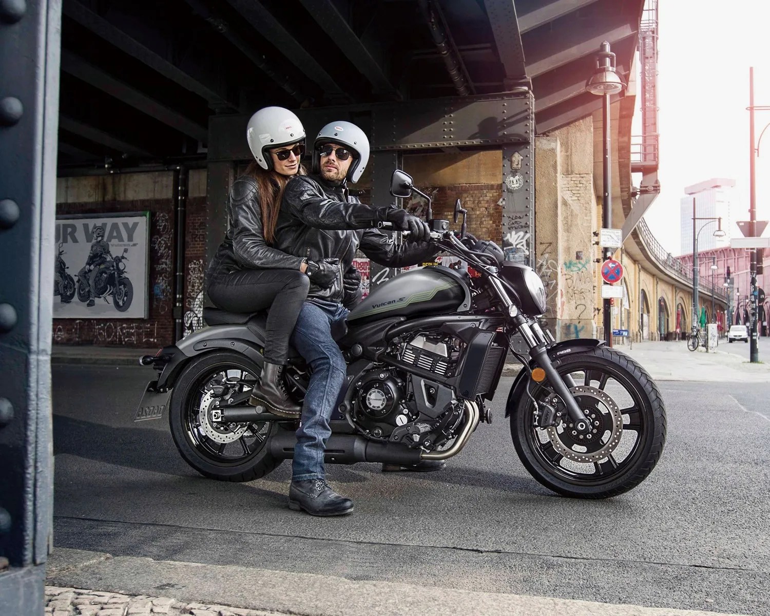 Black and gray Kawasaki Vulcan S motorcycle with two riders wearing white helmets and black leather jackets under a bridge.