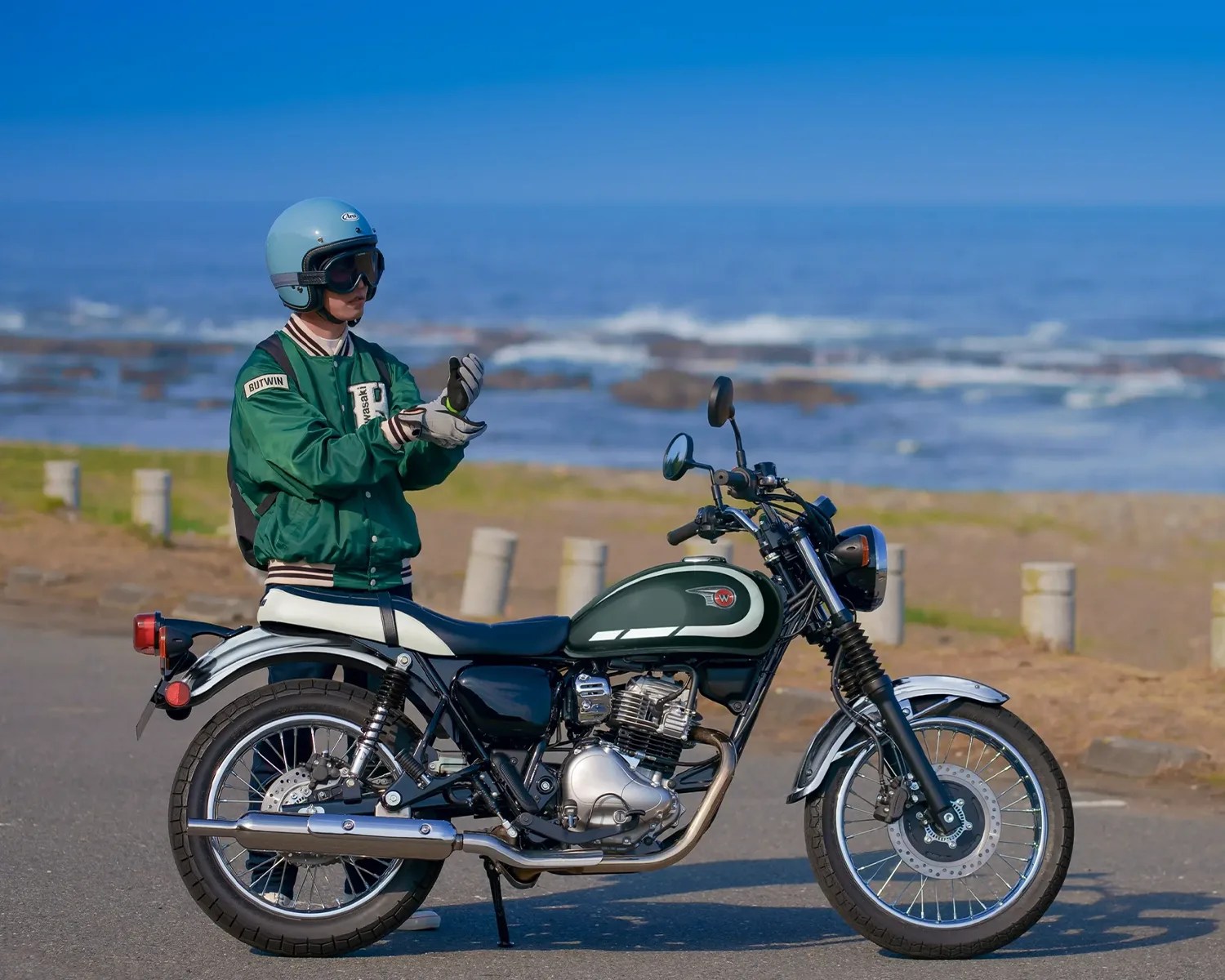 Person wearing a blue helmet and green jacket standing beside a green and white classic motorcycle near the ocean.