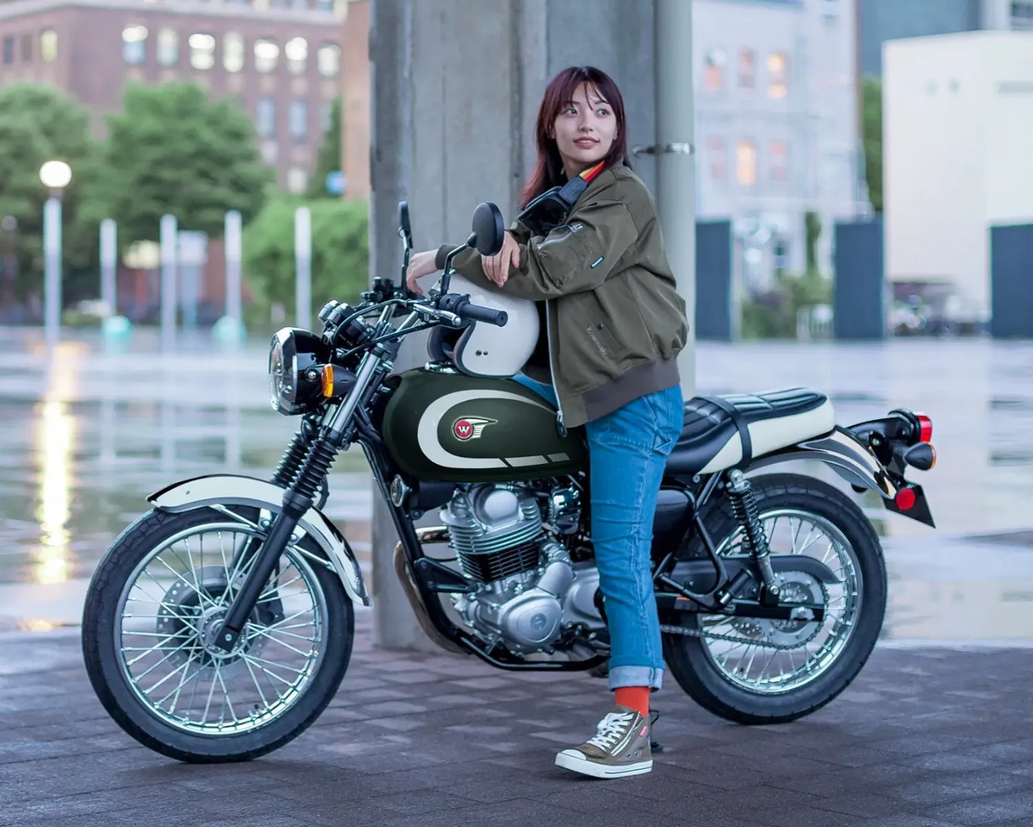 Person in an olive green jacket and blue jeans leaning on a green and white classic-style motorcycle.