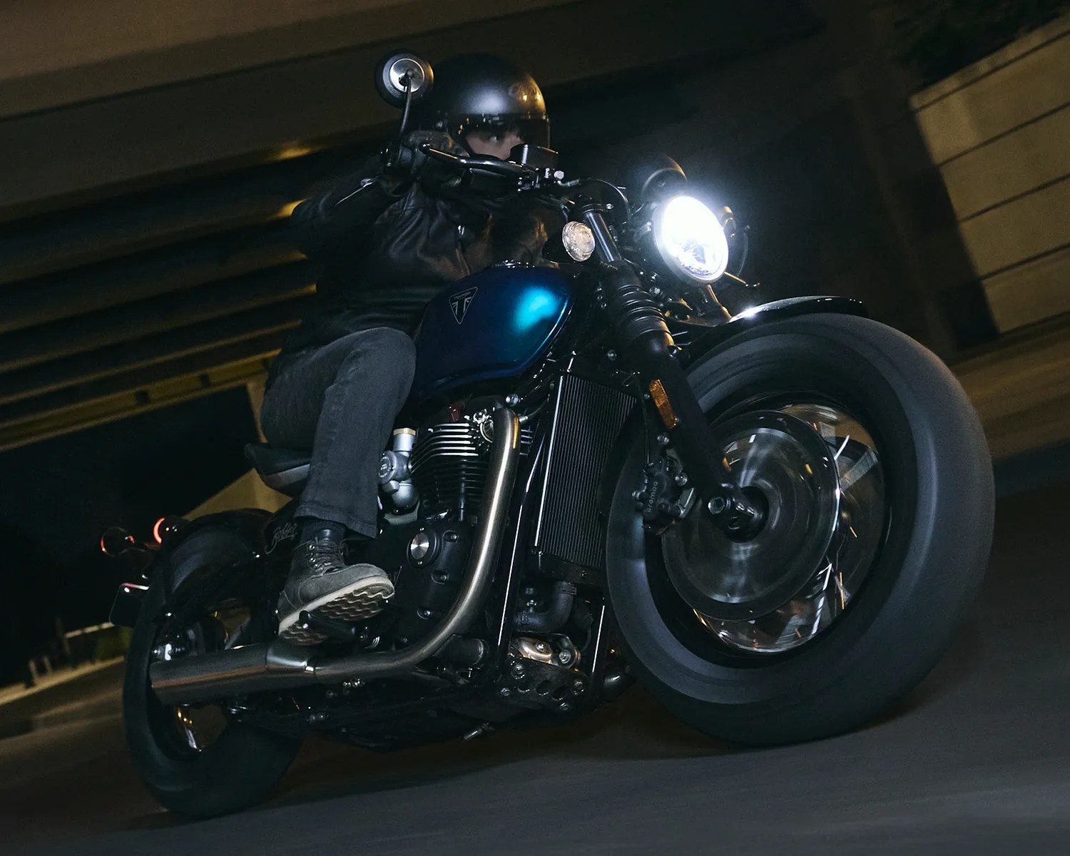 Person riding a blue Triumph motorcycle at night with the headlight on under a bridge.