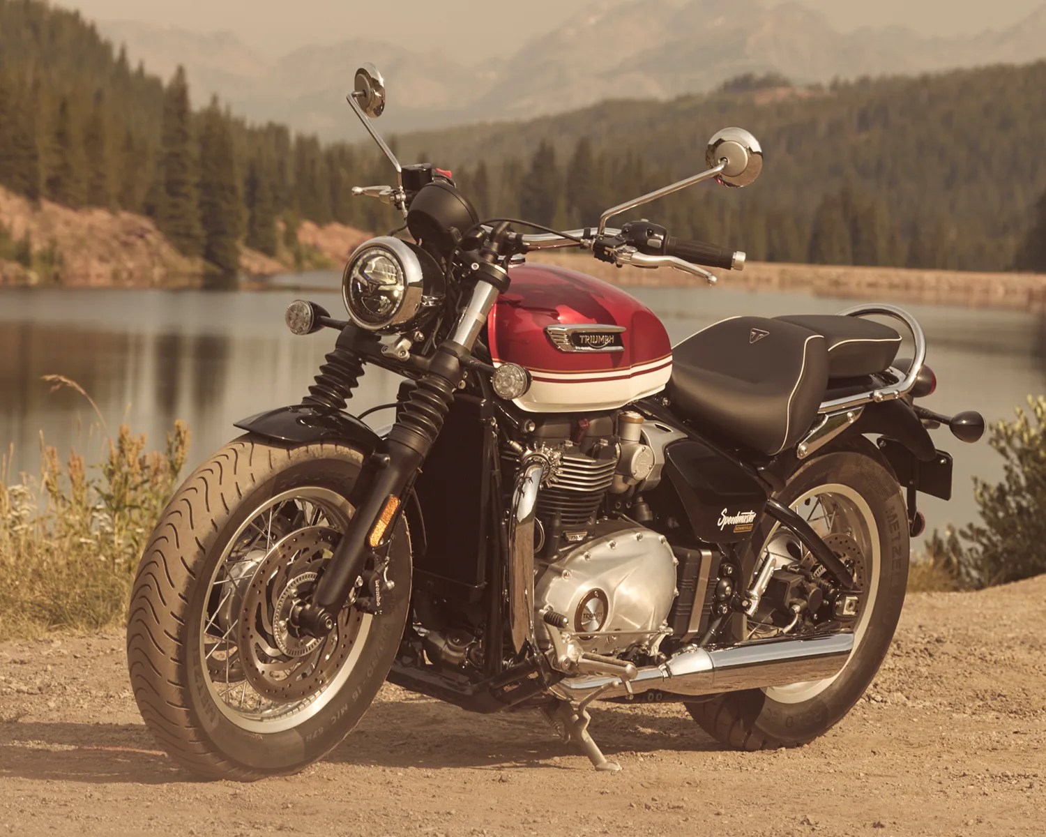 Red and white Triumph Speedmaster motorcycle parked on a dirt road near a lake with forested hills in the background.