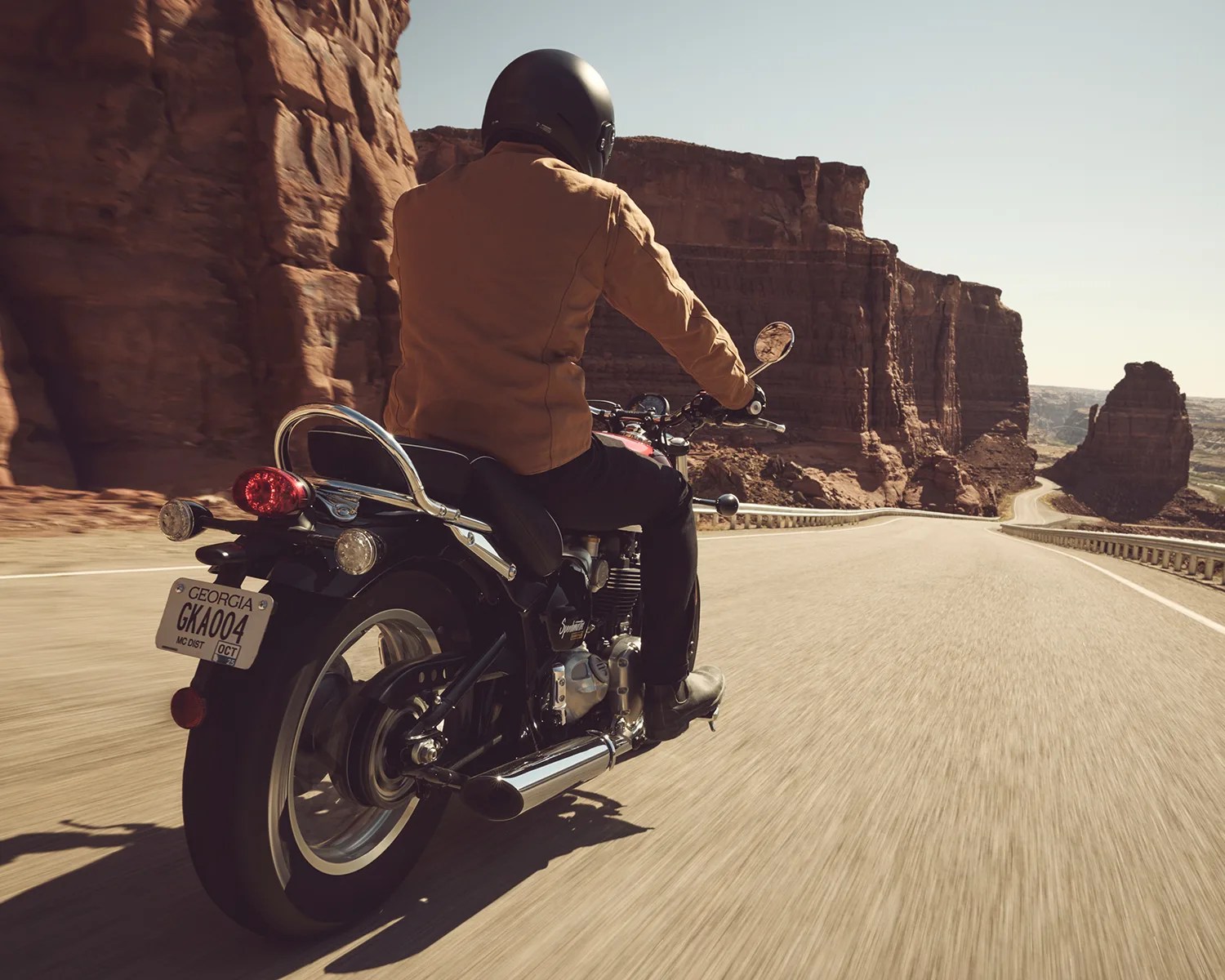 Person wearing a black helmet and brown jacket riding a black motorcycle on a desert road with red rock cliffs.