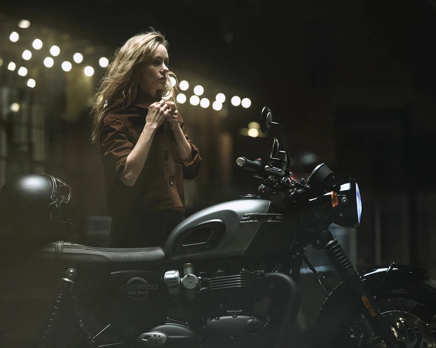 Woman in a brown jacket standing next to a black and silver Triumph Bonneville T120 Black motorcycle at night.